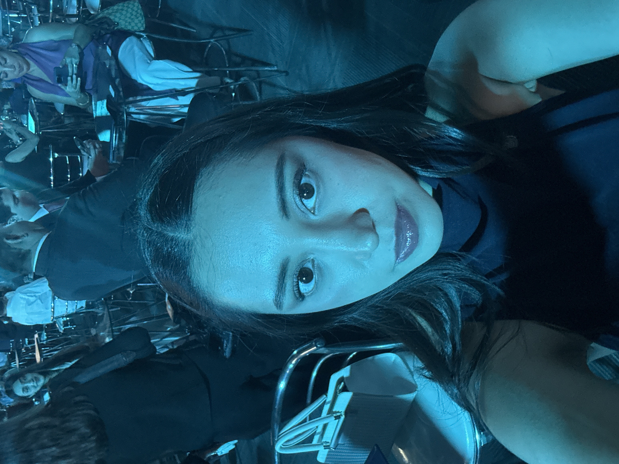 A medium-shot captures a woman with fair skin and dark hair, possibly of Latin American descent, in what appears to be a social gathering in Puebla, Mexico. She has dark eyes and is wearing lipstick. She is seated and slightly tilted her head, looking directly at the camera. Behind her, there are numerous people sitting on chairs, dressed in formal wear, some engaged in conversations. The lighting is a blue hue.