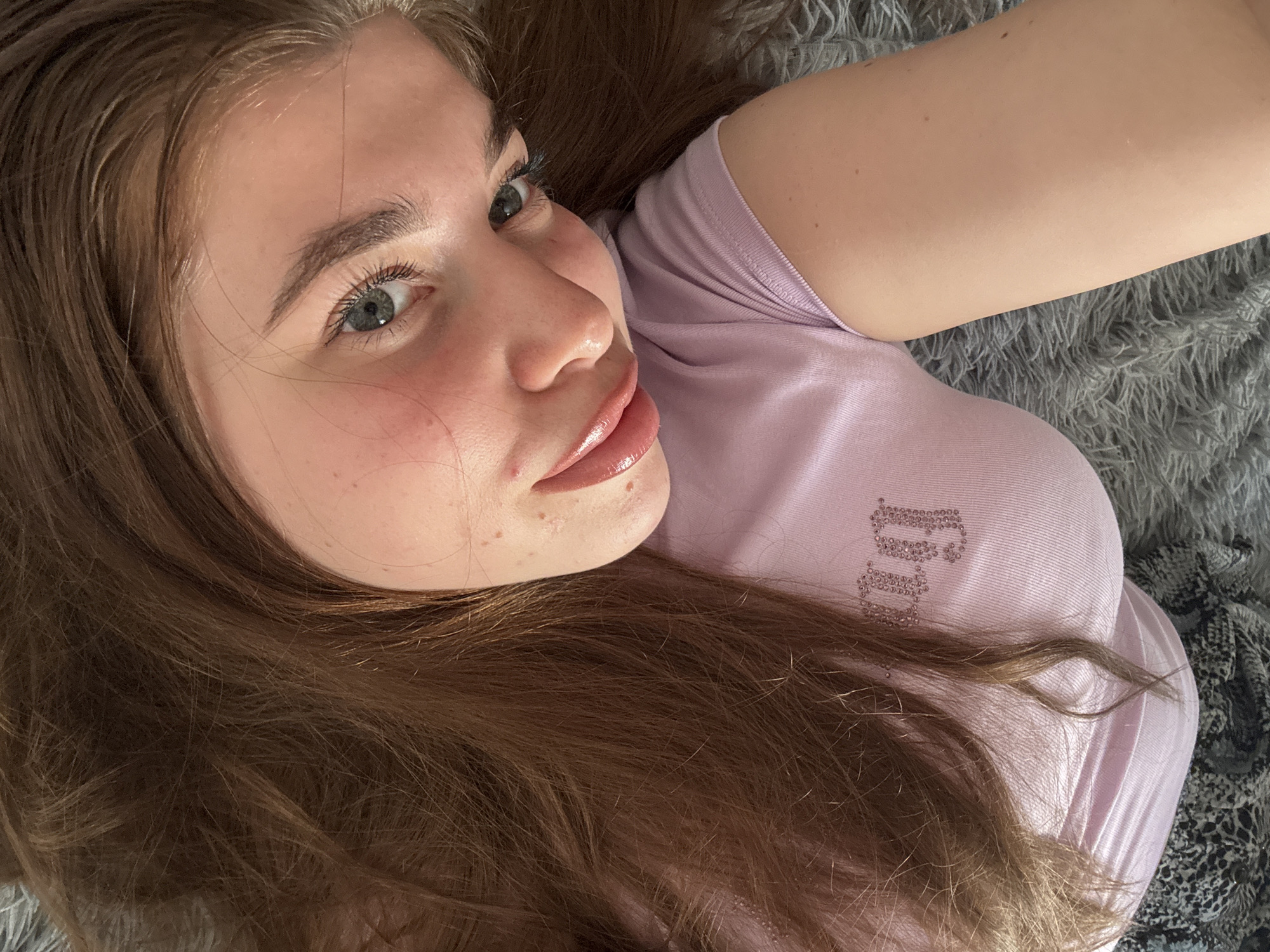 A young woman with long, brown hair lies on a soft, grey, furry surface, possibly a rug or blanket. She is looking up and to her left, with a slight smile and a playful expression. Her left eye is visible, showing a blue iris with flecks of grey. She is wearing a light purple t-shirt with sparkly, pink lettering that appears to say "JUICY" across the chest. The woman's skin is fair, with some light freckles visible on her cheeks and nose. Her lips are full and shiny, with a pinkish hue. The lighting suggests it is daytime, likely indoors, with natural light illuminating her face. The overall mood is relaxed and intimate. The location context provided is Мещёры, Russia.