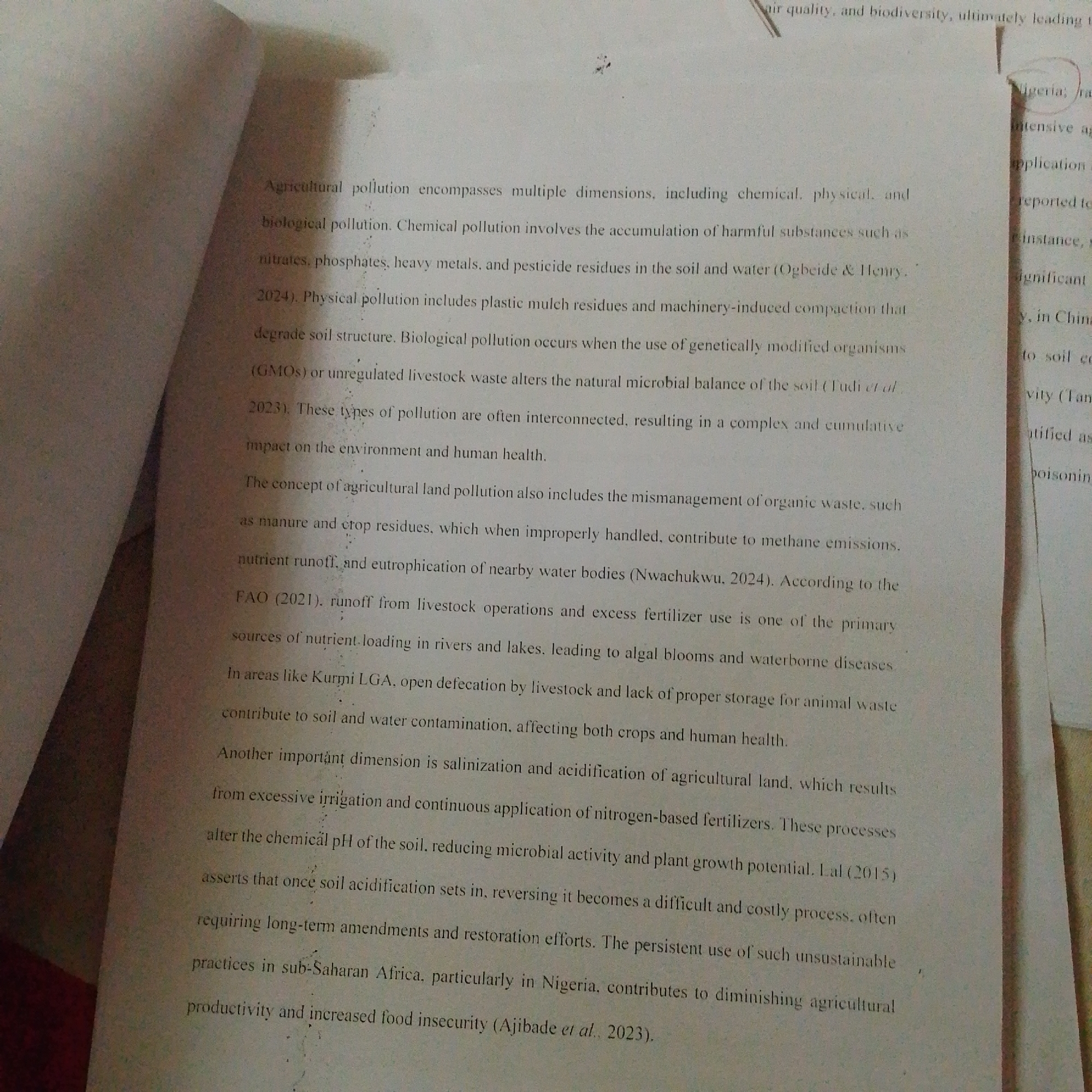 This is a photograph of text from a document. The text discusses agricultural pollution, its various forms (chemical, physical, biological), and its impacts on soil, water, and human health. It also mentions specific examples from Nigeria, including livestock operations in Kurmi LGA and unsustainable practices in sub-Saharan Africa. The text is printed on off-white paper and appears to be from a book or journal, with a folded page obscuring a portion of the text on the left side. There are no people, objects, or specific environmental details related to the city of Jalingo, Nigeria, present in the image.