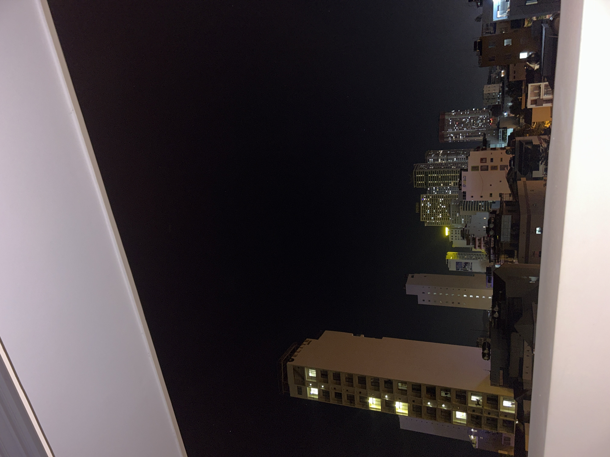 The image captures a nighttime view of an urban landscape, likely from an elevated perspective such as a balcony or high-rise building. The dominant feature is the vast, dark night sky, occupying the upper and central portions of the frame. Below and to the right, a dense collection of multi-story buildings illuminates the scene with countless glowing windows, suggesting a bustling city at night.
The buildings vary in height and architecture, appearing to be a mix of residential and commercial structures. Many windows in these buildings are brightly lit, casting a warm glow against the dark facades. In the mid-ground, a prominent building features a clear sign that reads "CO-OP Food" in English and Vietnamese characters, along with "CO.OPMART."
A broad, light-colored architectural structure, possibly a balcony railing or an adjacent wall, forms a strong diagonal element along the left side of the frame, partially framing the cityscape. The overall impression is one of a quiet, clear night in a densely populated area. The scene depicts an ordinary evening in Bắc Nha Trang, Vietnam, with no specific activity or events taking place beyond the ambient life of the city.