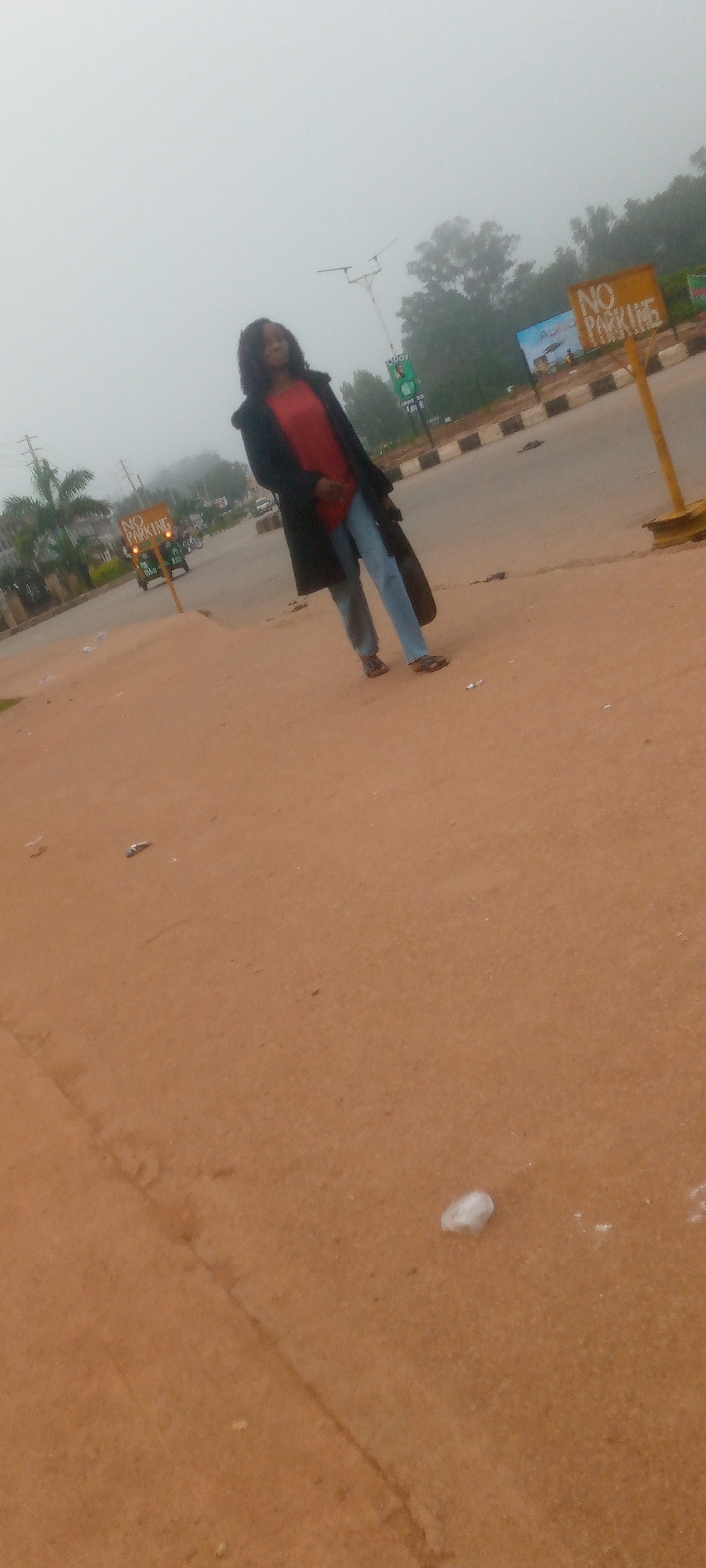 A woman stands on a dusty road, looking towards the camera. She is wearing a black coat over a red shirt and blue jeans, with sandals on her feet. A large black bag is slung over her left shoulder. In the background, a yellow "NO PARKING" sign is visible, along with a street light and trees. The sky is overcast and foggy. The scene appears to be in Gura Top, Nigeria.