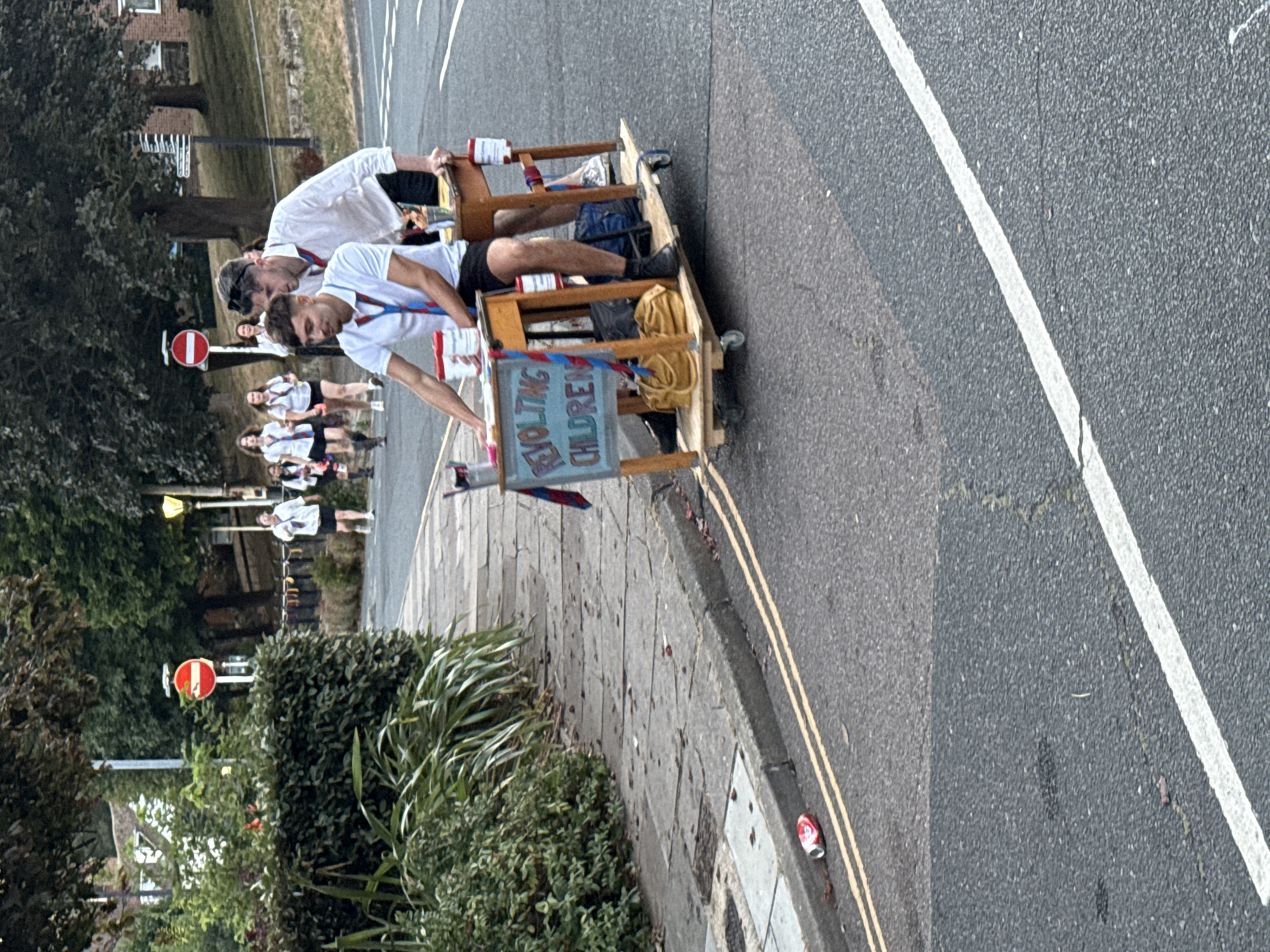 Two young males, dressed in school uniforms consisting of white shirts and dark shorts, are positioned on a makeshift wooden cart with wheels. The cart is supporting two small desks, one in front of the other. The front desk has a sign attached that reads "REVOLTING CHILDREN" in colorful lettering. The individuals appear to be pushing or riding the cart along a paved road. In the background, several other individuals, also in school uniforms, are walking on a sidewalk. The environment includes trees, a sidewalk, a road with yellow double lines, and a red and white circular traffic sign. A discarded red and white can is visible on the side of the road. The scene is consistent with a school event or celebration.