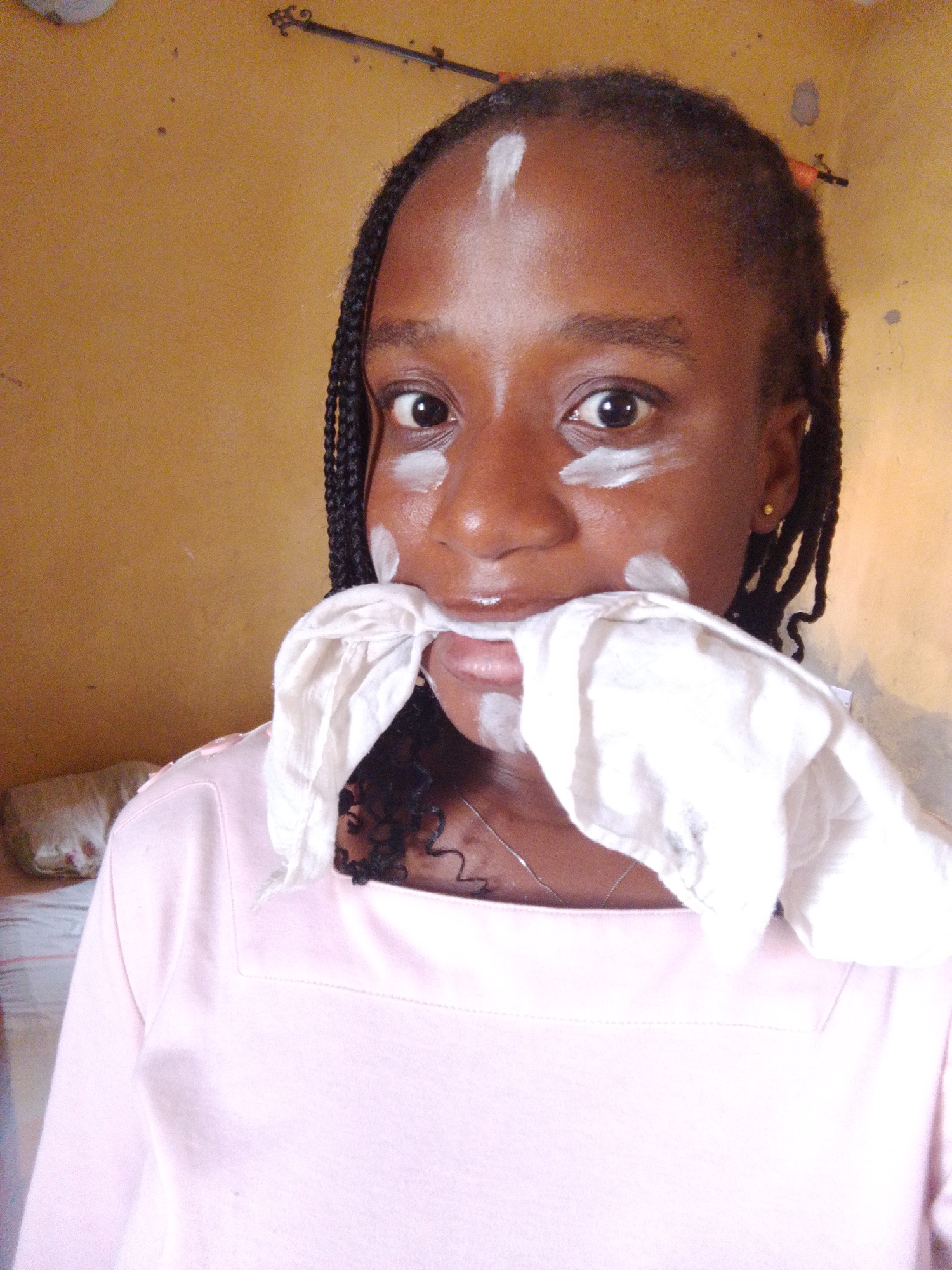 The image shows a close-up of a young Black woman, likely a teenager, with dark braided hair. She is wearing a light pink, square-necked top and a silver chain necklace. White cream or makeup is applied in streaks on her forehead, under her eyes, and on her cheeks, and a white cloth is held in her mouth, obscuring her lips. She is looking directly at the camera. The background is a textured, yellowish-orange wall, common in residential areas in Umuahia, Nigeria. The lighting suggests an indoor setting, possibly a bedroom or living area, with a hint of a bed and pillow visible in the blurred background on the left. The overall scene appears to be a candid, informal moment captured by a self-taken photograph.