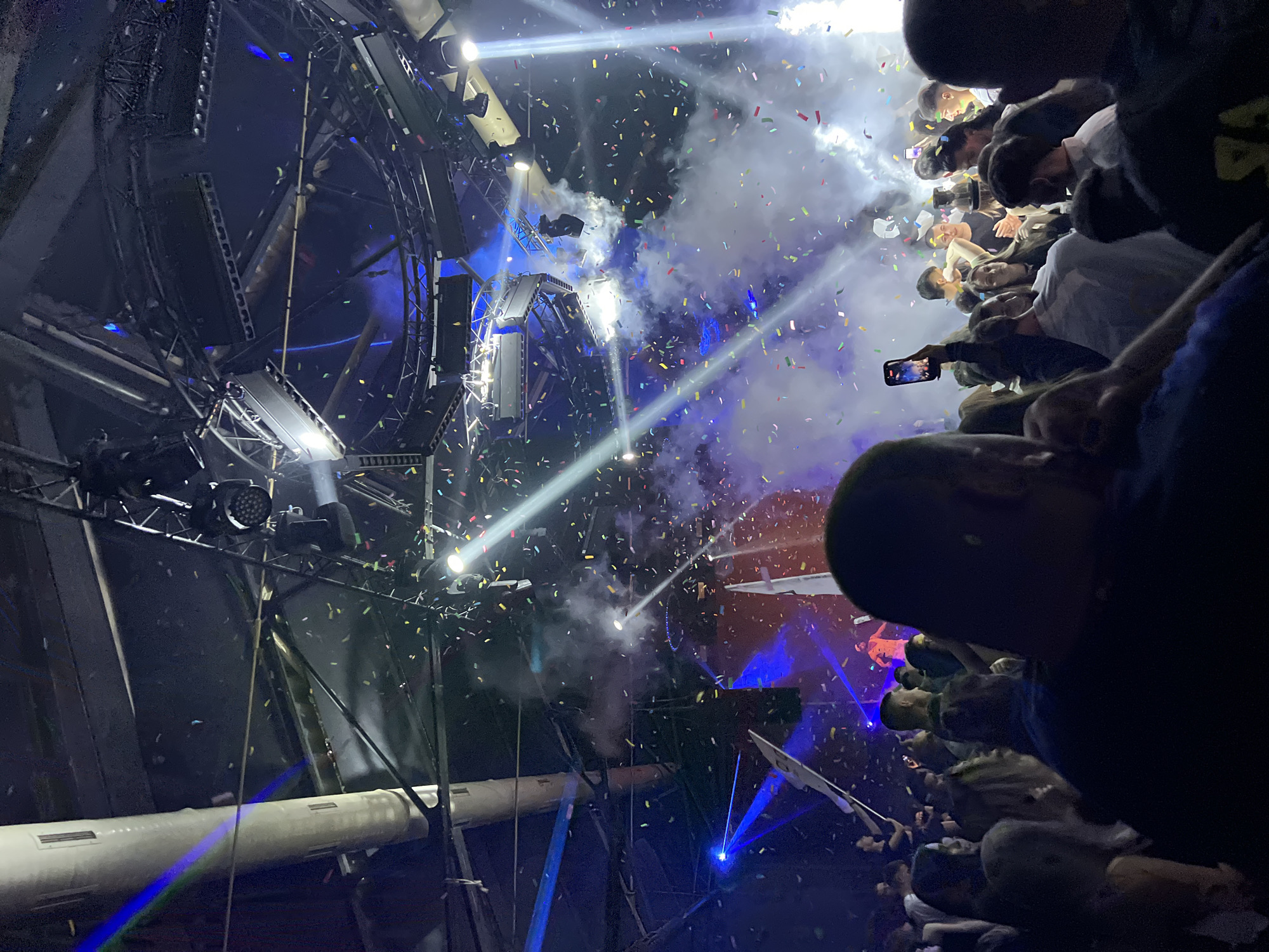 A low-angle view captures a concert atmosphere within a venue, presumably located in Burgas, Bulgaria. The image is filled with stage lighting illuminating falling confetti and plumes of smoke. A large, complex structure of sound equipment and lighting rigs dominates the left side of the frame. On the right side, a crowd of people are visible, with many holding up phones to record the event. The overall impression is one of a vibrant, energetic concert experience.