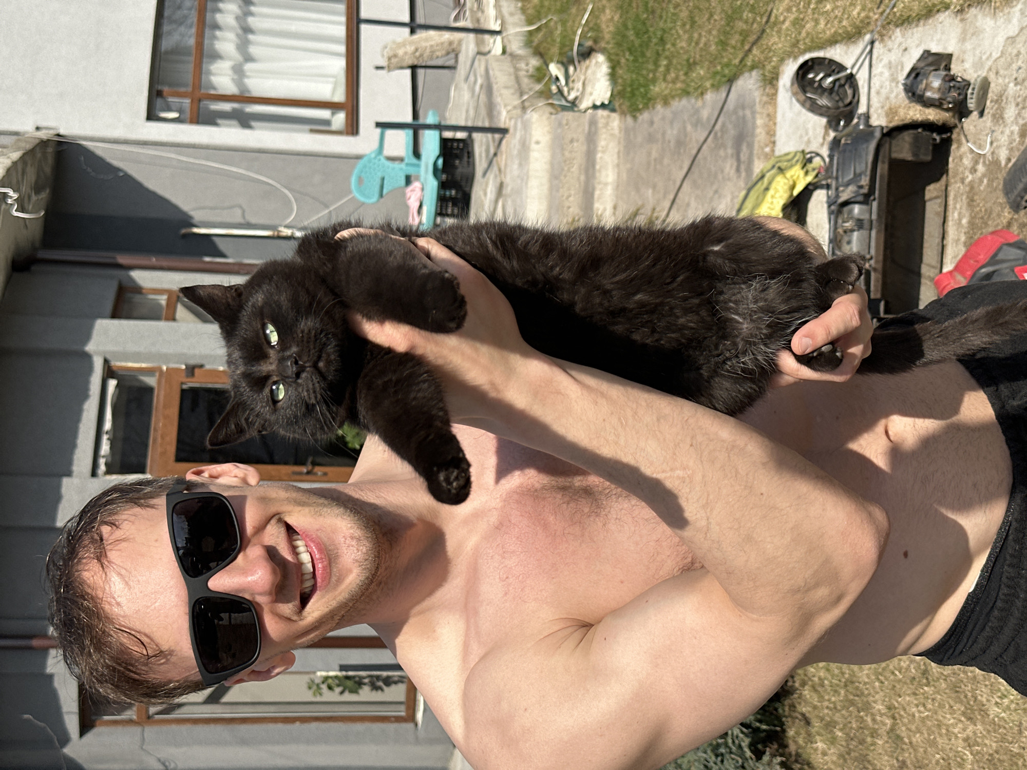 A man wearing sunglasses holds a black cat in an outdoor setting. The man is shirtless and smiling. The background includes a building with windows, various objects including a chair and what appear to be machine parts. The location is Kursk, Russia.