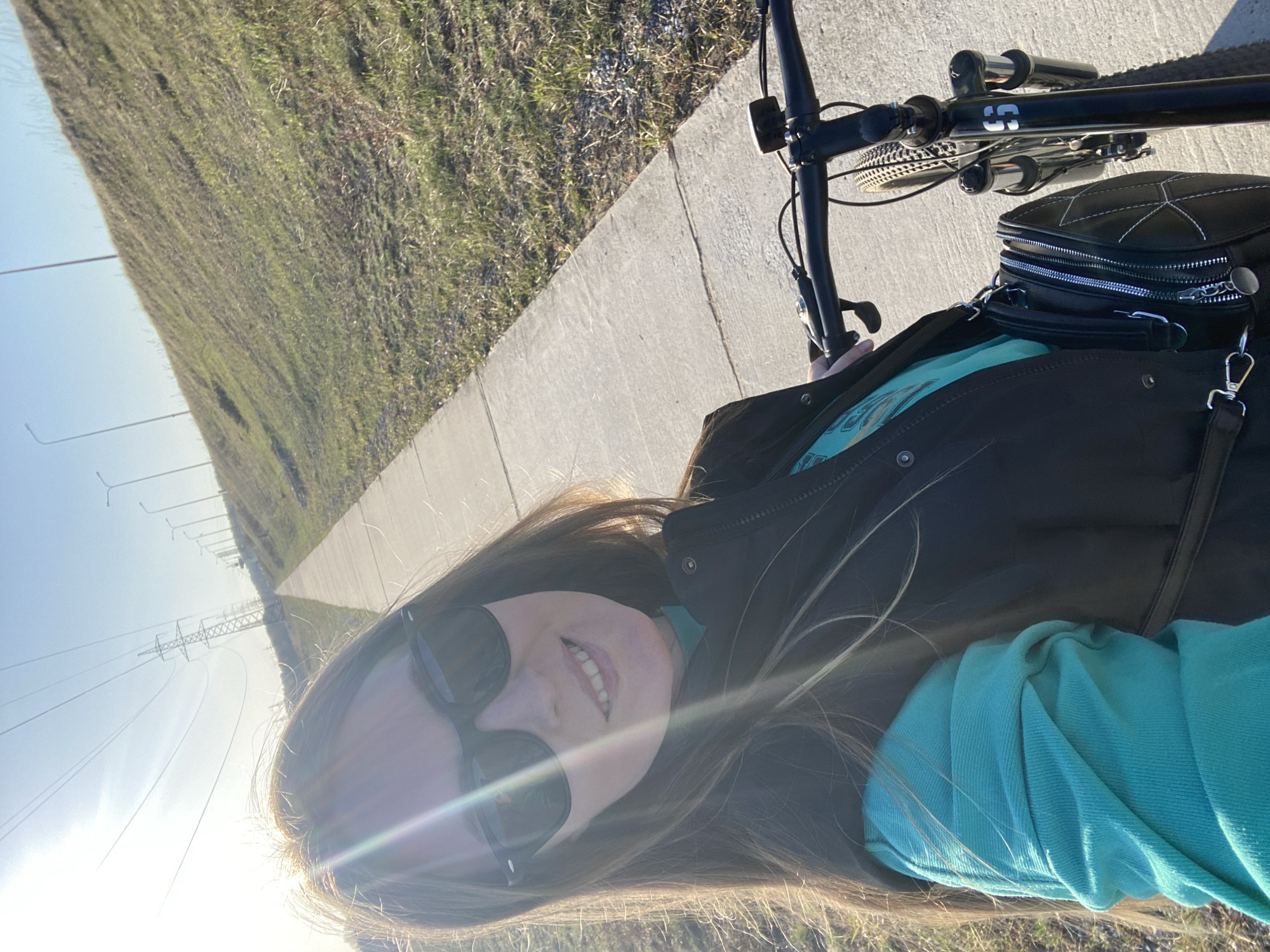 A woman wearing sunglasses and a black vest over a turquoise shirt is taking a selfie while riding a bicycle. The bicycle has a black frame and is equipped with a black bag. She is riding on a concrete path next to a grassy area. In the distance, there are utility poles and power lines. The bright sunlight indicates it is daytime.
The location is determined to be the city of Chaikovsky, Russia, based on the presence of power lines and a concrete path next to a grassy area, which is consistent with infrastructure in the region.