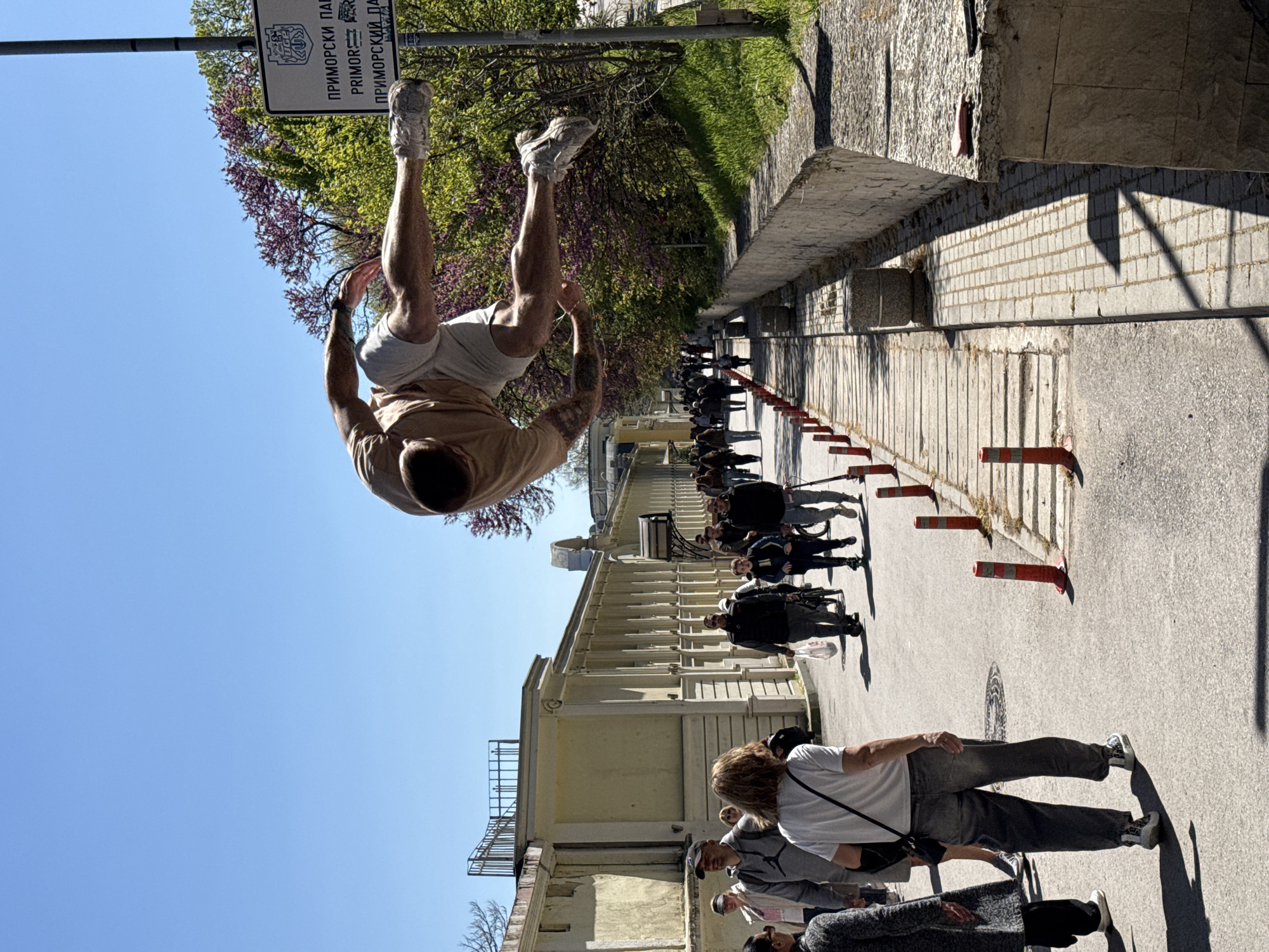 A street-level image in Varna, Bulgaria, shows a man in mid-air, performing a backflip. He is wearing athletic shoes, light-colored shorts, and a tan t-shirt. His legs are nearly vertical, and his head is near the ground. Visible tattoos adorn his arms.
In the background, a line of people walks along a sidewalk bordered by orange and white traffic pylons. A beige building runs parallel to the sidewalk. A signpost with Bulgarian text is partially visible above the man. The sky is clear and blue.