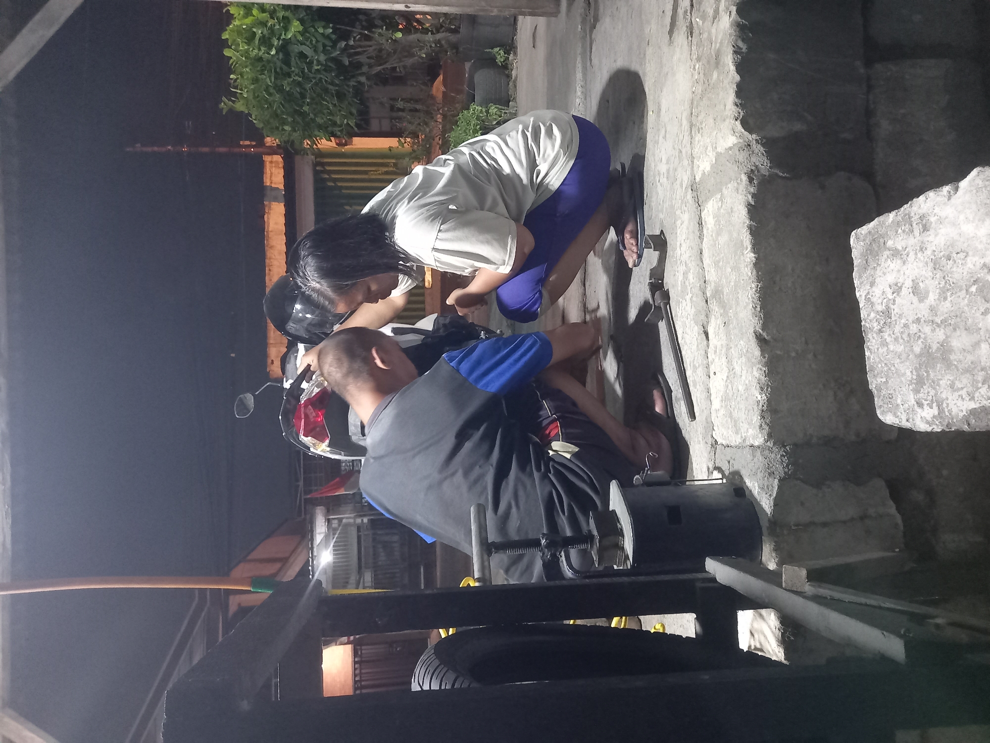 In the city of Ikerikergeger, Indonesia, two individuals, a man and a woman, are crouched down next to a motorcycle. The man, wearing a black t-shirt with blue accents and dark shorts, is holding a tool, possibly for tire repair. The woman, dressed in a light-colored t-shirt and purple leggings, is also focused on the area in front of the motorcycle. Adjacent to them is a motorcycle, partially visible, with a black helmet on its seat. The scene appears to be outdoors, possibly a residential area or a workshop, with a concrete ground surface and some overgrown vegetation in the background. The lighting suggests it is evening or night. Tools and equipment are scattered around, indicating a repair or maintenance activity.