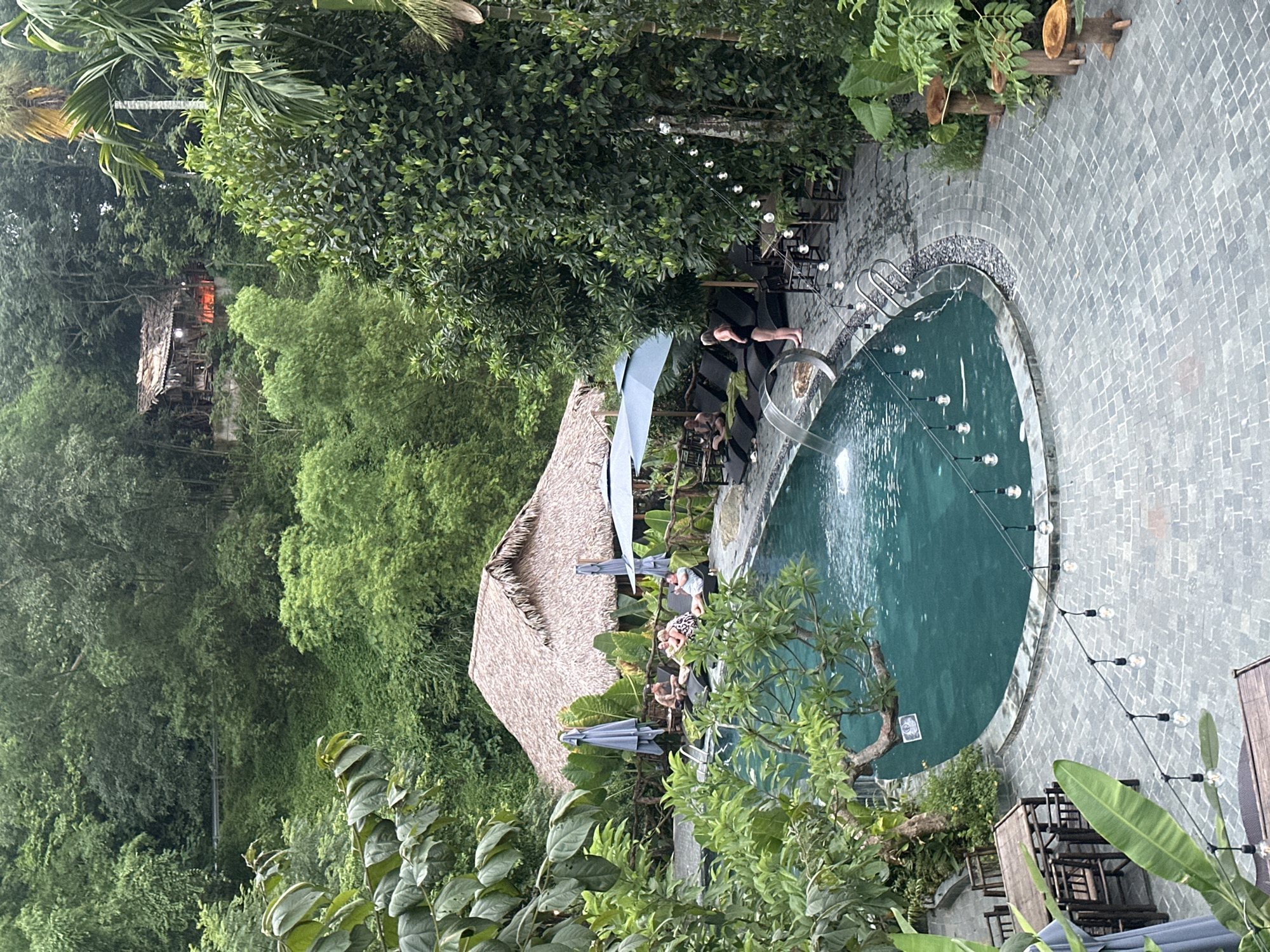 An aerial view shows a resort area in Pù Luông Commune, Vietnam. A crescent-shaped swimming pool with dark teal water is the focal point, featuring a cascading water feature and a ladder. String lights with exposed bulbs hang over the pool and the adjacent paved area. Several people are relaxing on lounge chairs around the pool, some under umbrellas. A thatched-roof building, possibly a restaurant or common area, is visible behind the pool. The entire scene is nestled within a lush, green environment with dense trees and foliage surrounding the resort structures. In the distance, on a hillside, a rustic wooden building with a corrugated roof is visible, with an orange glow emanating from within. The ground is paved with grey interlocking stones. The overall atmosphere suggests a tranquil, nature-oriented retreat.