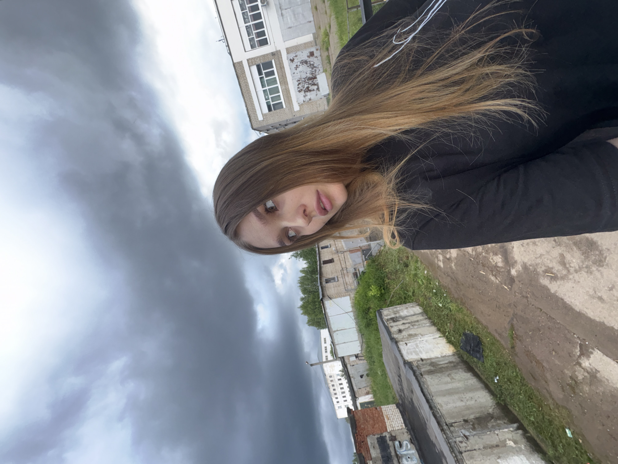 A young woman with long, blonde-brown hair and brown eyes is captured in a selfie taken outdoors. She is wearing a black long-sleeved shirt. The background suggests an industrial or utilitarian area, possibly within the city of Chaikovsky, Russia. Several brick and concrete structures, resembling garages or storage units, are visible. Some of these structures show signs of wear and tear, with graffiti and peeling paint. Overcast, dark clouds dominate the sky, hinting at impending rain or a stormy weather condition. The ground appears to be a dirt or gravel path, with patches of grass and scattered debris. The overall scene conveys a somewhat somber or moody atmosphere due to the weather and the utilitarian surroundings.