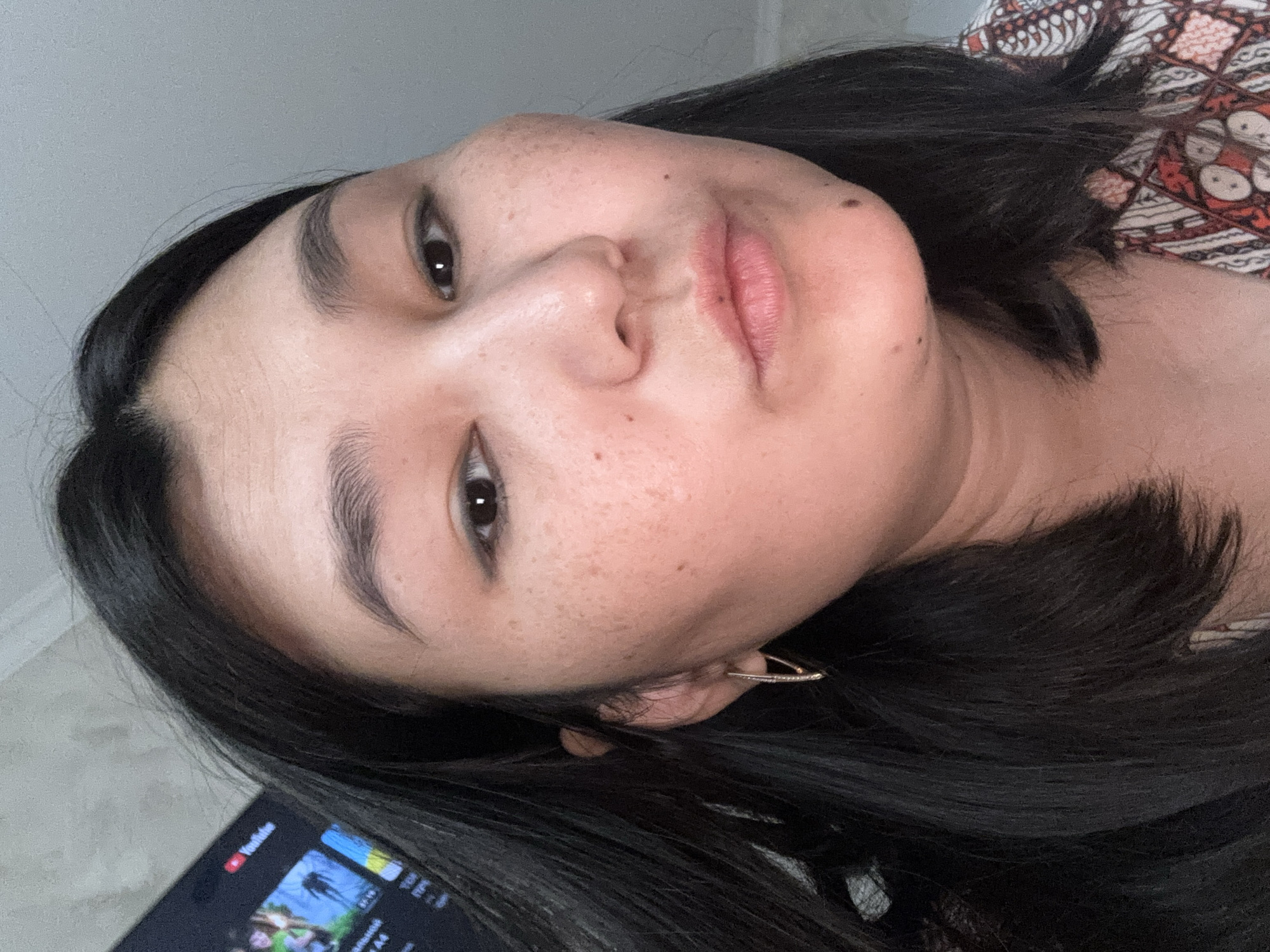 This is a close-up selfie of a woman in the city of Kyzylorda, Kazakhstan. She has dark hair, dark eyes, and fair skin with freckles. She is wearing a patterned shirt. In the background, a television screen is visible, displaying the YouTube logo. The overall environment appears to be indoors, likely a home.