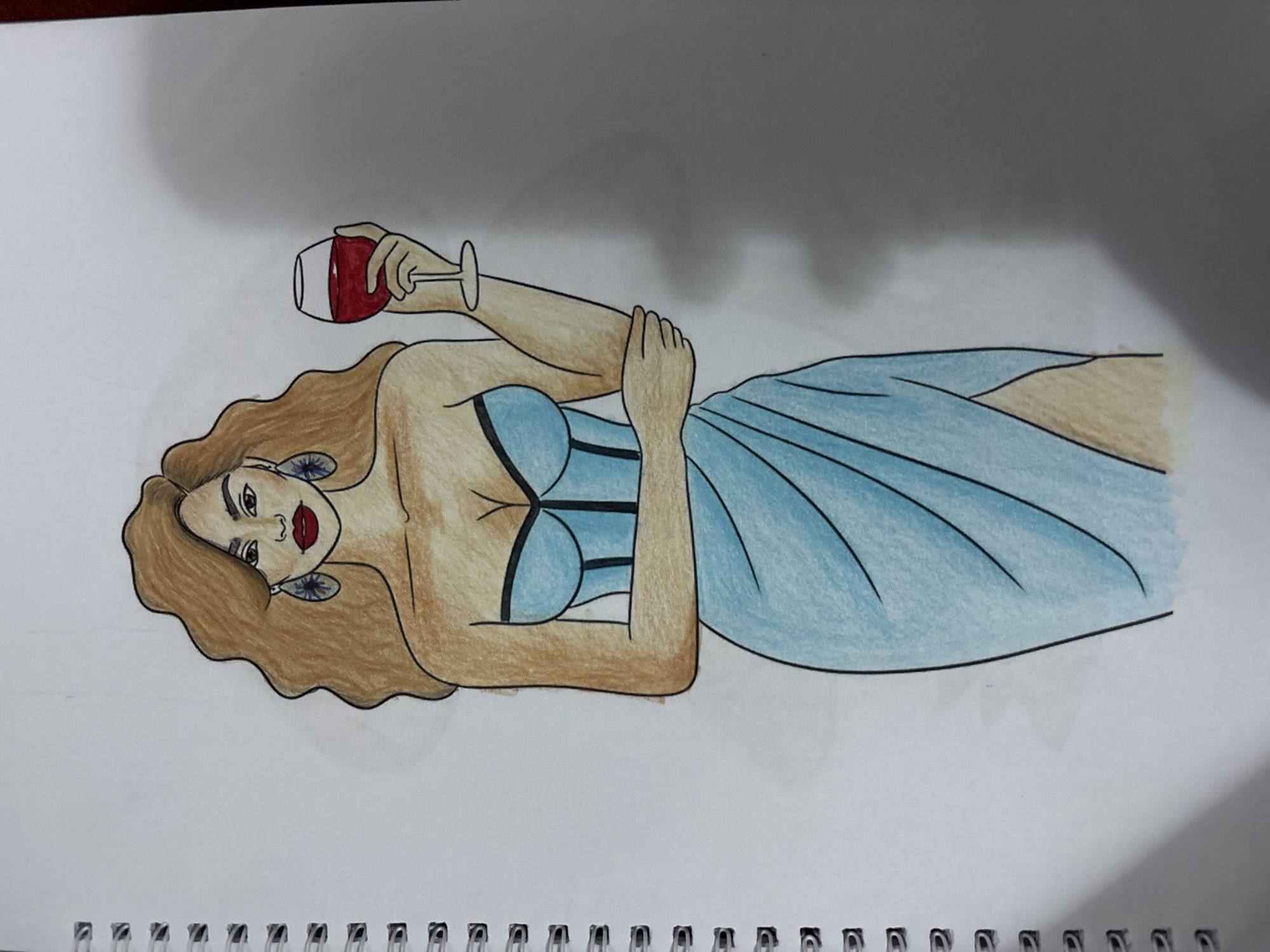The image is a drawing on a spiral-bound notebook page, depicting a woman with long, wavy brown hair, fair skin, and bold red lipstick. She is wearing large, ornate blue earrings and a light blue, strapless dress with a high slit. The dress has black detailing around the bust. She is holding a stemmed wine glass filled with red liquid in her right hand. Her left arm is bent, with her hand resting on her hip. The background is white, with a soft gray shadow cast to the right of the drawing. The notebook's spiral binding is visible on the left side of the image. There is no indication of a specific location in Lafia, Nigeria, or any urban environment. The drawing is a fashion illustration.