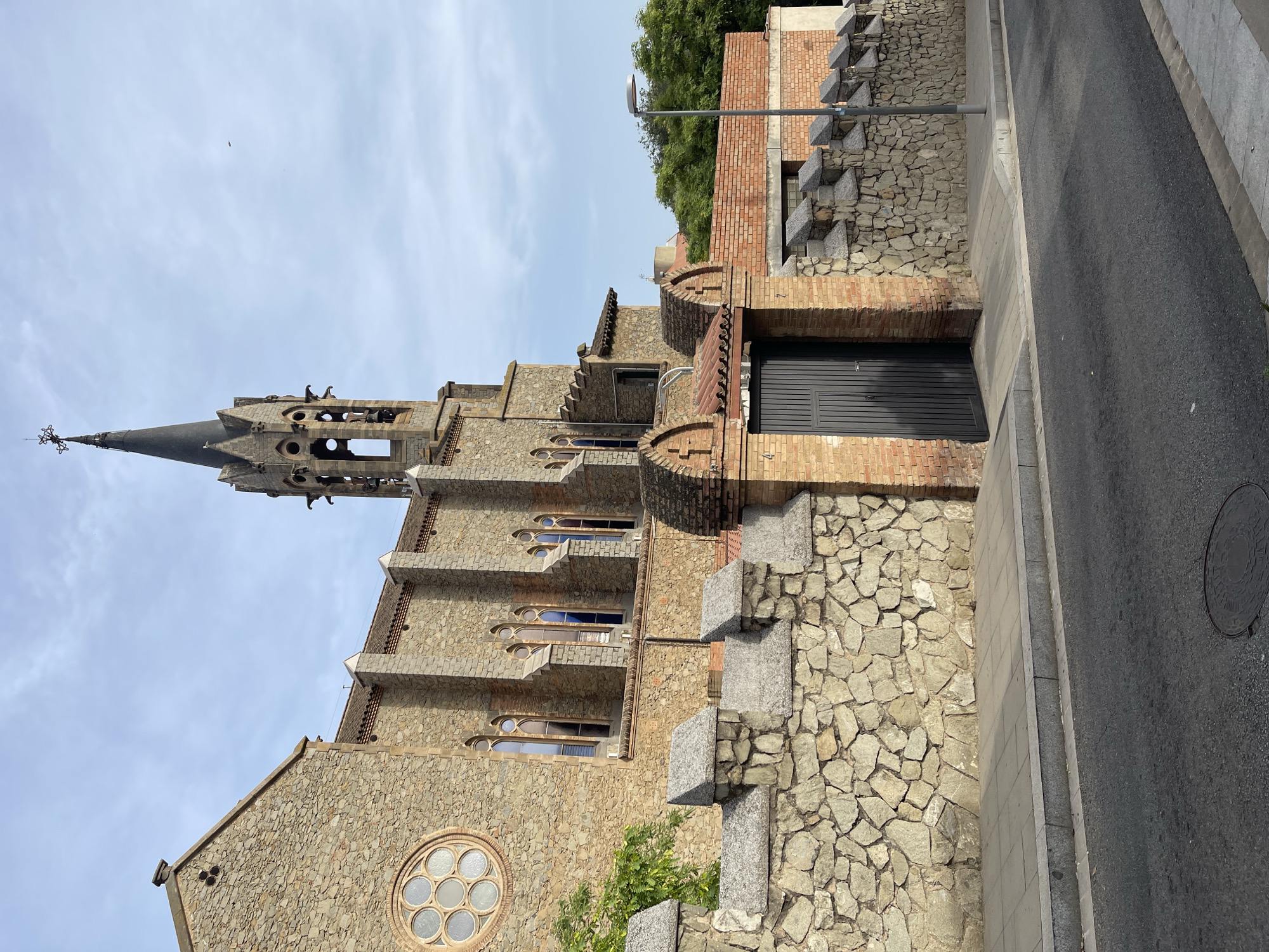 The image shows the exterior of a stone church building with a tall, pointed bell tower. The church has several gothic-style arched windows and a circular rose window on the left side. A stone wall with crenellated battlements runs along the front of the property, separating it from a paved sidewalk and an asphalt street. A dark gray metal gate is set within a brick section of the wall. Green trees are visible behind the wall and to the right of the church. The sky is partly cloudy. The building is identified as the Església de Sant Francesc d'Assís in Santa Coloma de Gramenet, Spain.
