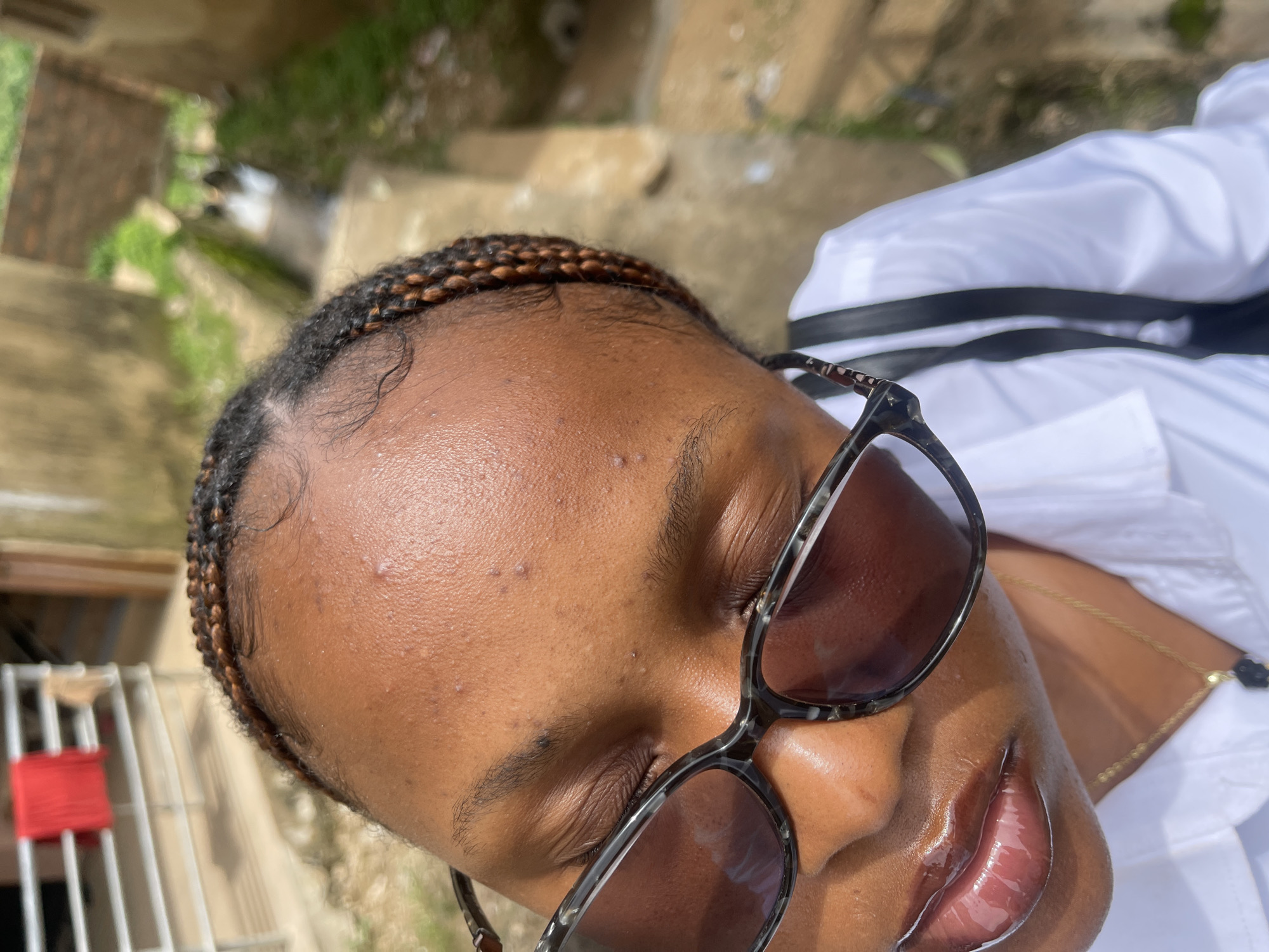In Rafin Par, Nigeria, a young woman with braided hair and a white collared shirt is captured in a close-up, angled shot. She is wearing dark-rimmed glasses and has glossy lips. A thin gold necklace with a dark pendant is visible around her neck. In the background, a red cloth is draped over a metal railing, and a narrow, somewhat overgrown drainage ditch runs alongside a textured, light-colored wall. The overall environment suggests an outdoor urban setting.