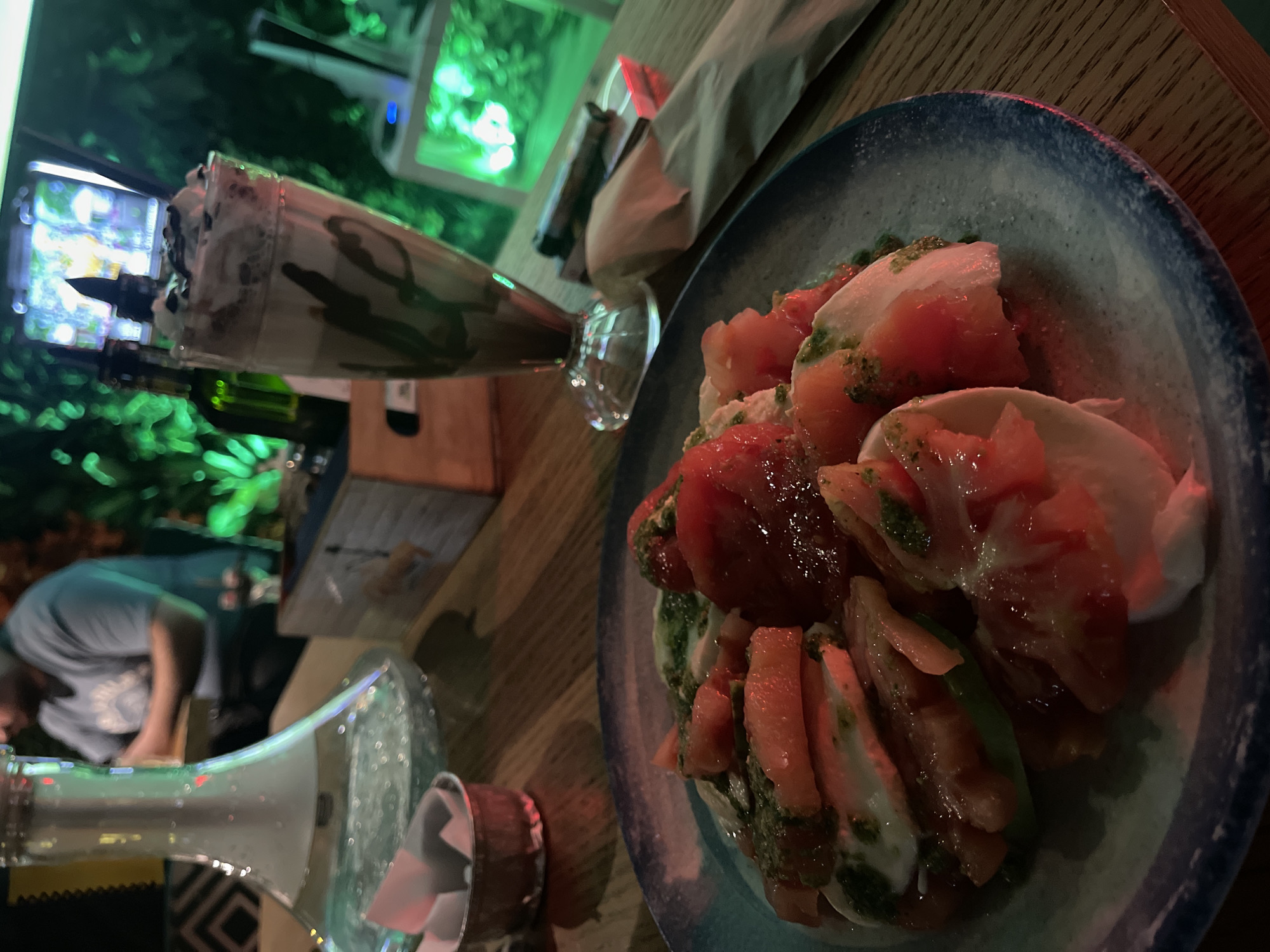The photograph depicts a table setting indoors, likely at a restaurant or bar. A plate of sliced tomatoes and mozzarella, drizzled with pesto, is prominently featured. To the left, a tall glass filled with a milkshake-like beverage topped with whipped cream and chocolate shavings is visible. In the background, indistinct figures of people are present. A glass bottle and what appears to be a container are placed near the edge of the table. The setting is illuminated by ambient lighting. There is no definitive evidence to confirm that the location is in Sofia, Bulgaria.