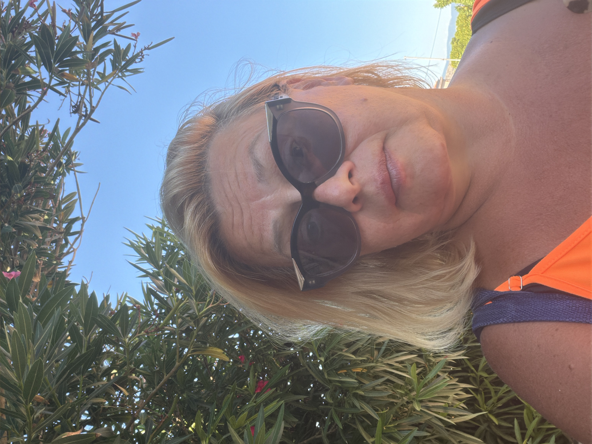 This is a close-up, outdoor, selfie-style photograph of a fair-skinned woman with blonde hair and sunglasses, taken on a sunny day. She is wearing an orange top. Behind her, there is lush green foliage and a clear blue sky. The location is identified as Općina Baška, Croatia.