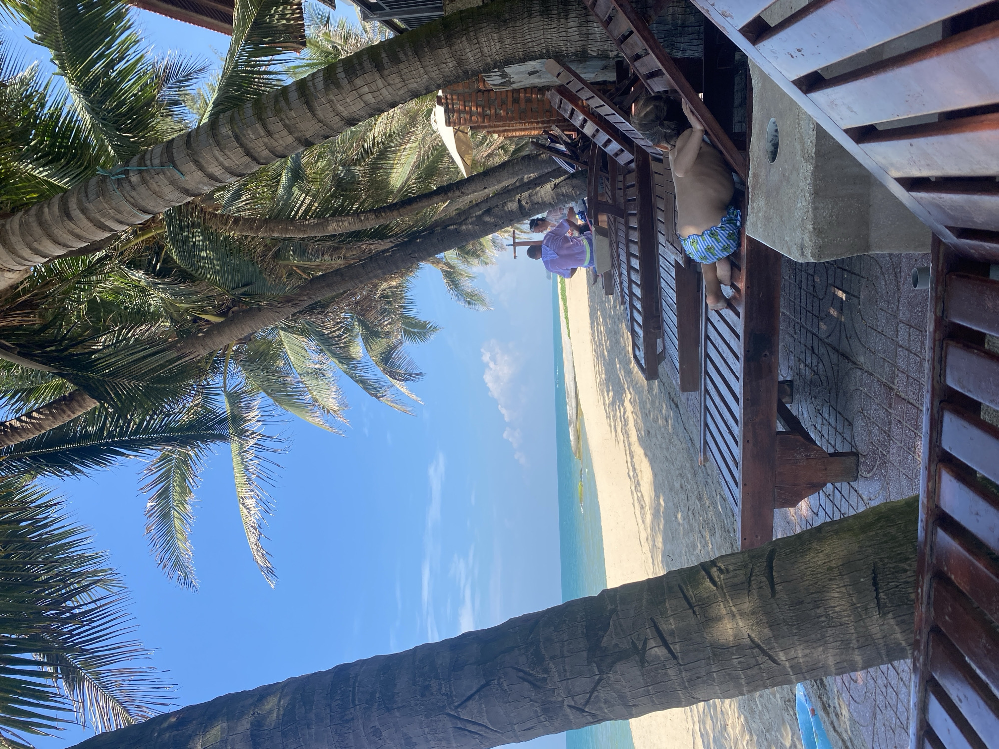 The image depicts a tropical beach scene, likely in Phu Thuy Ward, Vietnam, under a clear blue sky. In the foreground, several wooden lounge chairs are arranged on a tiled patio area, partially shaded by the fronds of tall palm trees. A young child, wearing patterned swim trunks, is lying face down on one of the lounge chairs, appearing to be resting or playing. Further back, two individuals, a man in a purple shirt and a woman in a white top, are seated on lounge chairs, looking towards the ocean. The background features a wide expanse of sandy beach, transitioning to the turquoise waters of the sea. The overall atmosphere suggests a relaxed vacation setting.