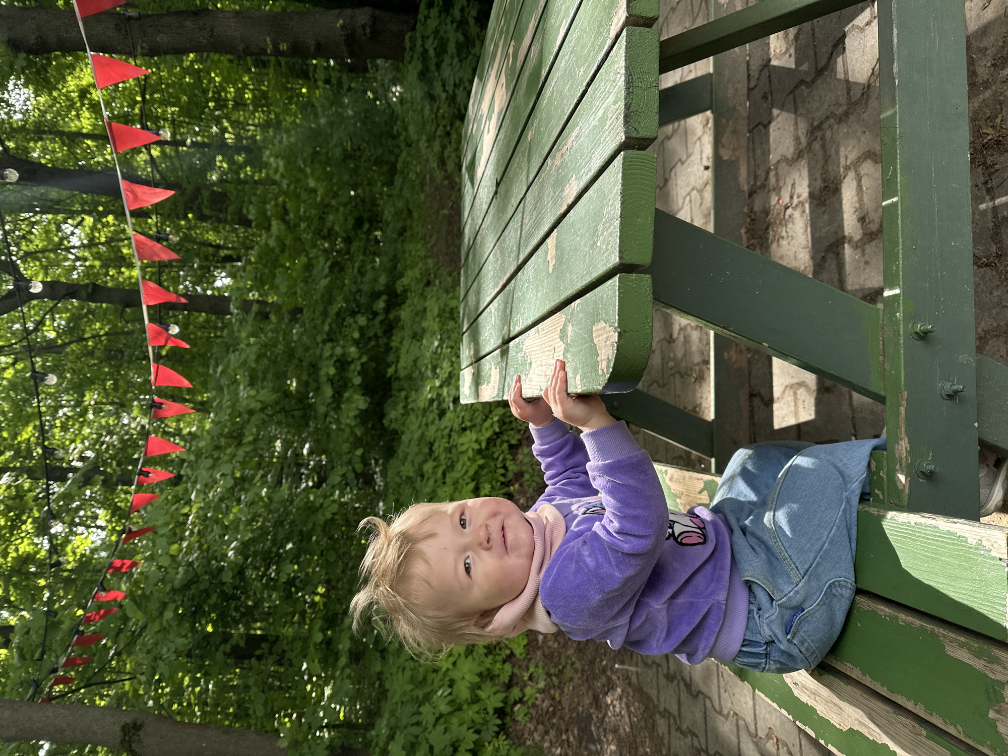 A young child wearing a purple sweatshirt and jeans is holding onto the edge of a green picnic table. The picnic table is located outdoors on a paved surface near a wooded area with trees and a string of red triangular flags hanging above. The location appears to be a park or recreational area. The precise location within the city of Nizhny Novgorod, Russia, cannot be ascertained from the image.