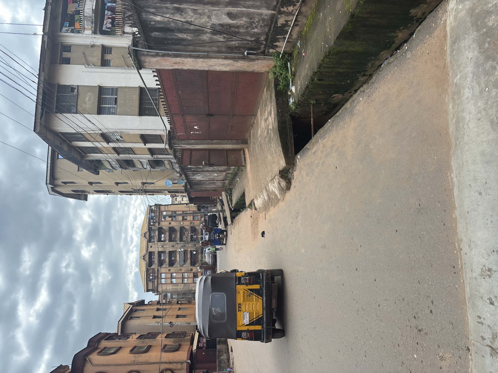 This is a street-level view of a residential area in Onitsha, Nigeria, under a cloudy sky. In the foreground, a yellow and black auto-rickshaw (also known as a Keke Napep) is parked on the right side of the asphalt road, facing away from the viewer. The road is worn and uneven in places, with a concrete gutter running along the right side, partially overgrown with moss and containing some litter.
Further down the street, two individuals are visible. One appears to be standing near a blue motorcycle laden with goods, and another person is walking further ahead on the right side of the road. The environment is characterized by multi-story buildings with balconies and exposed electrical wires crisscrossing overhead. A large, rusty red metal gate is prominent on the right side, part of a wall that runs along the street. The buildings exhibit signs of wear and tear, with some areas of peeling paint. The overall impression is of a densely populated urban environment.