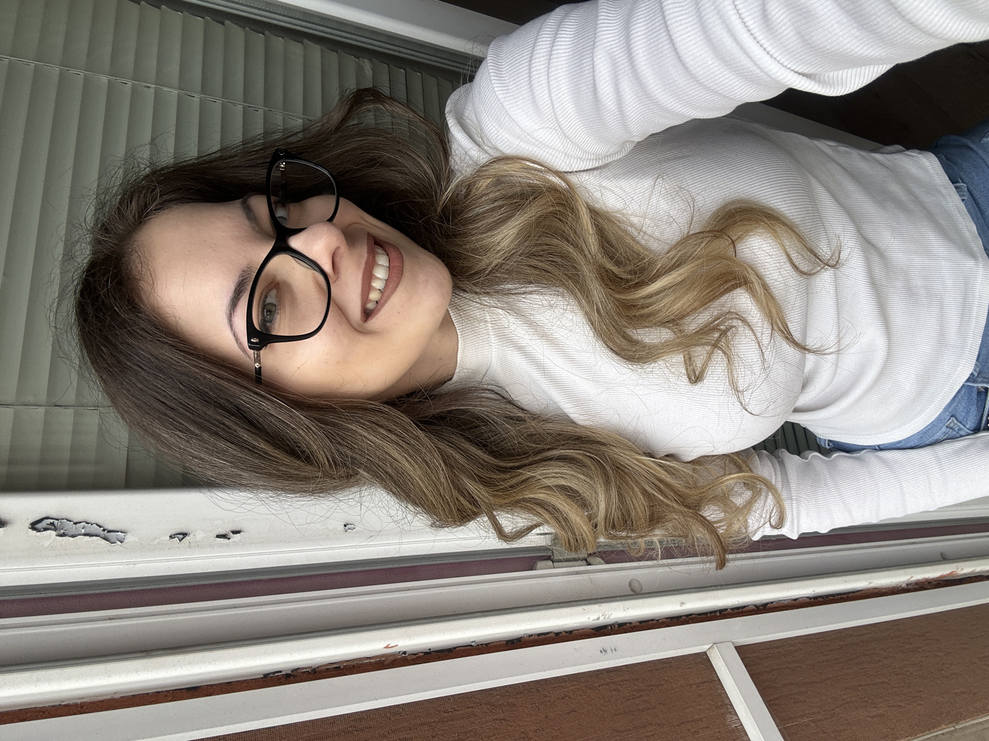 A woman with long, wavy hair that transitions from dark brown to blonde is smiling and looking directly at the camera. She is wearing black-framed glasses and a white, long-sleeved ribbed shirt. She appears to be indoors, with window blinds visible behind her. The bottom of the image shows the frame of a window, with peeling paint and a screen. There is no evidence of the city of Stara Zagora, Bulgaria in the image.