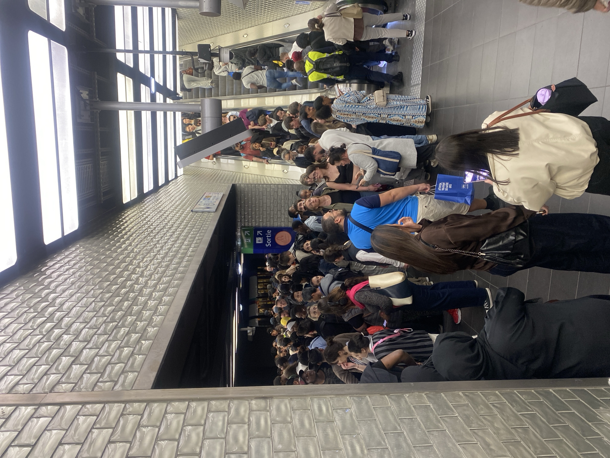 The media file captures a densely crowded underground passage or metro station, viewed from a slightly elevated perspective, looking down and across the multitude of people. The image is rotated 90 degrees counter-clockwise.
The primary subjects are a large group of people, standing shoulder-to-shoulder, filling the width of the passage. They appear to be queuing or moving slowly towards an exit or escalators, which are visible further in the background. Many individuals are engaged with their mobile phones. Clothing styles vary, from casual wear to more formal attire, and several people are carrying bags or backpacks. A person wearing a bright yellow vest is noticeable near the escalators.
The setting is an indoor, artificially lit environment. The walls in the foreground and along the passage feature distinct white, glossy rectangular tiles, characteristic of some Parisian metro stations. Bright, elongated light panels illuminate the ceiling and upper walls.
Notable details include a blue and green sign with an arrow pointing right and the word "Sortie," which is French for "Exit," strongly indicating the location is in France, specifically Paris as per the context. Another visible object is a blue shopping bag with text "Festival de Guerre" and "GAP." The time of day is not discernible due to the indoor lighting. The atmosphere appears to be one of patient waiting despite the significant crowd density. Escalators are visible at the far end of the passage, with more people ascending.