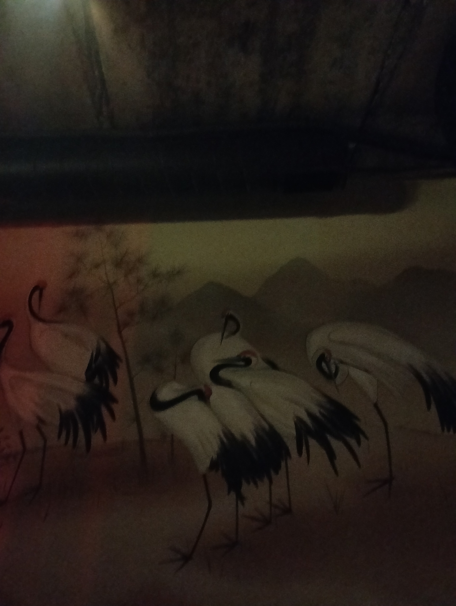 The image shows a decorative mural or wallpaper depicting a group of cranes in a traditional East Asian style. The background features soft, indistinct mountains. Above the mural is a dark, solid shape, possibly a structural element like a beam or pipe, that is out of focus. The exact location in Ivano-Frankivsk, Ukraine is not apparent from the image. There are no people visible.