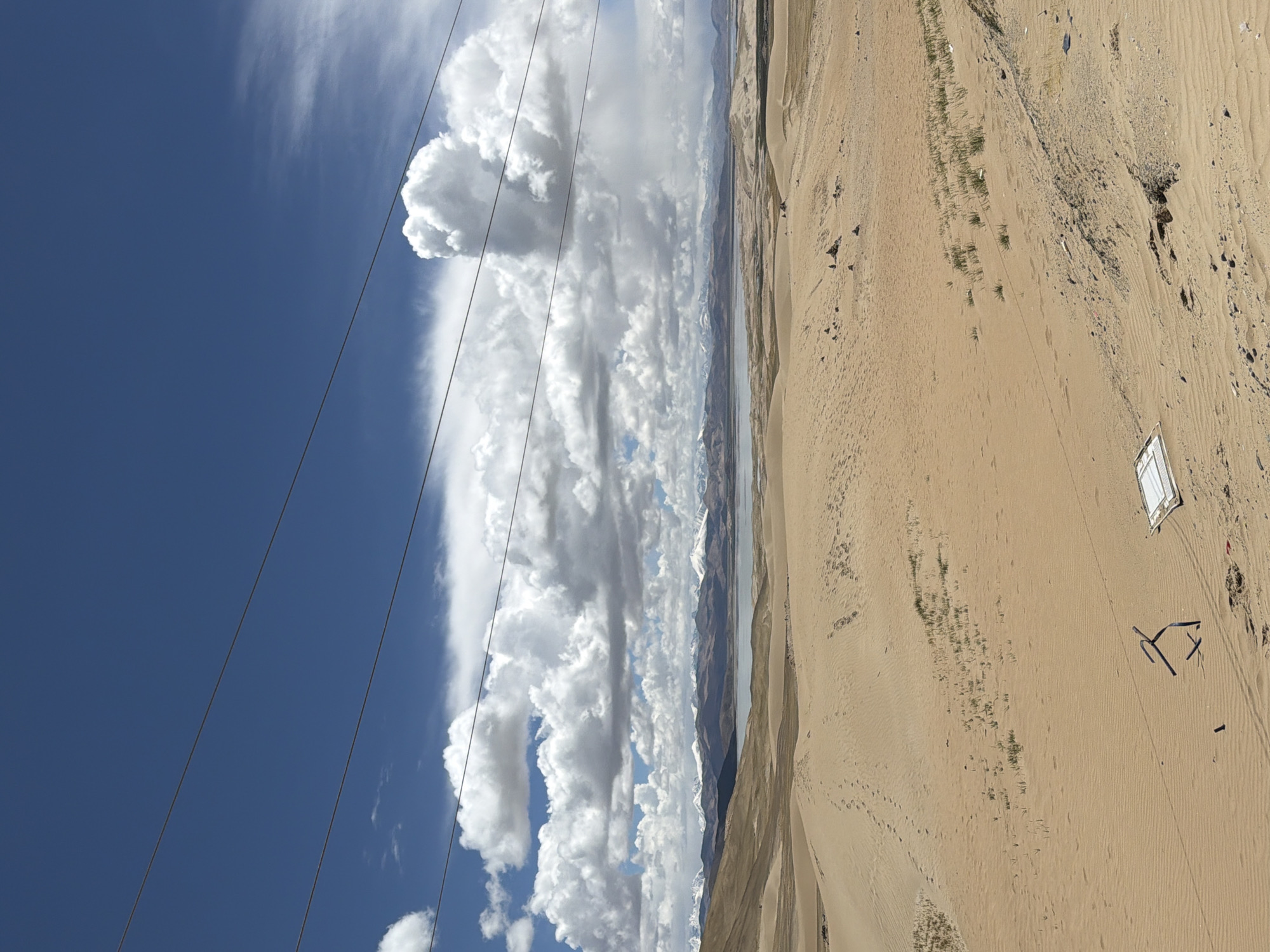 The image captures a vast, arid landscape under a dramatic sky, likely in Labrang township, China. The scene is dominated by a sweeping expanse of sandy desert terrain in the foreground and midground, characterized by undulating dunes, ripples in the sand, and scattered patches of sparse, greenish vegetation. Visible within the sandy area are numerous tire tracks, suggesting recent vehicle movement, and footprints.
In the distance, a narrow body of water, possibly a lake or river, stretches horizontally, separating the desert from a range of formidable mountains. These mountains are partly covered with snow on their peaks, indicating a high-altitude environment. The sky above is a brilliant blue, filled with an impressive array of large, white cumulus clouds that stretch across the horizon, casting shadows and creating varied textures. Several power lines, black and thin, traverse the upper left portion of the sky, running diagonally across the frame.
On the sandy ground in the lower right, there's a small, white rectangular object, resembling a canvas or a makeshift shelter. Nearby, some dark, irregularly shaped debris, possibly metal or plastic, lies scattered. The time of day appears to be bright daylight, with clear visibility. No people are visible, and no specific activity is taking place, although the tire tracks imply past human presence. No visible text can be discerned. The overall impression is one of immense scale, natural beauty, and isolation, with subtle hints of human interaction with the environment.