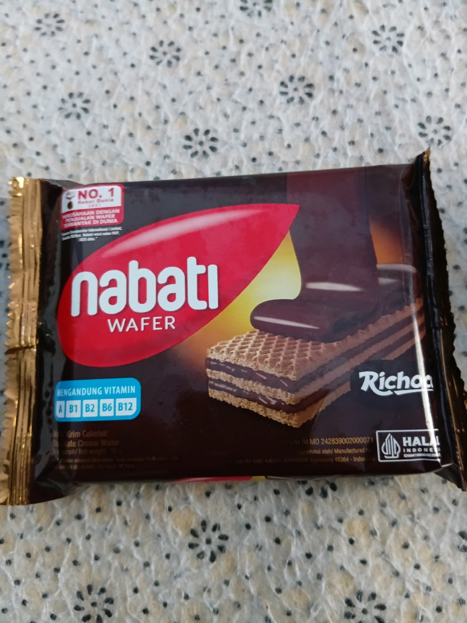 The image shows a close-up of a Nabati Wafer Richoco product packaging. The packaging is primarily dark brown with a red oval shape in the center displaying the Nabati logo in white lettering. Below the logo, it says "WAFER" in smaller white letters. To the left of the oval, there is a red banner with "NO. 1 Rekor Dunia 1921" printed in white. Below this, it states "PERUSAHAAN DENGAN PENJUALAN WAFER TERBANYAK DI DUNIA" which translates to "Company with the most wafer sales in the world." At the bottom of the red oval, there's a blue rectangle that says "MENGANDUNG VITAMIN" (Contains Vitamins) with vitamin labels A, B1, B2, B6, and B12. The image also features a visual representation of a wafer biscuit with chocolate filling, and the word "Richoco" is printed in white script on the right side of the packaging. A HALAL logo is visible on the bottom right. The product is placed on a textured, light-colored surface with a repeating pattern of small black dots and scattered floral-like designs. The background surface appears to be a textile or a textured placemat. There are no people or animals visible in the image. The lighting is even, casting minimal shadows. The image quality is good, with clear focus on the product packaging. No specific location within Tanggul Wetan, Indonesia is discernible from the image itself.
