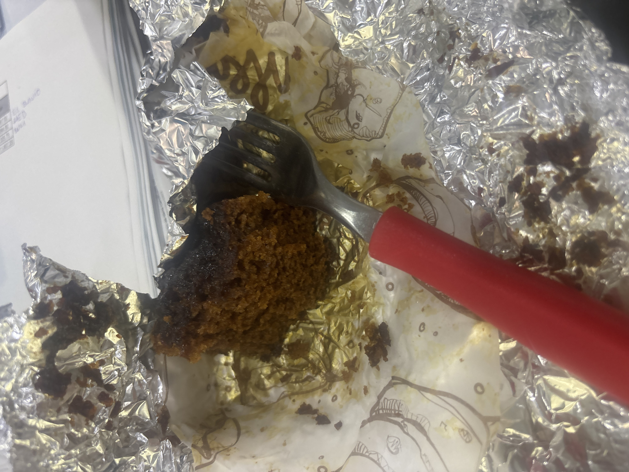 The image shows a close-up view of a partially eaten dark brown cake or muffin, presented in a white paper liner and wrapped in crumpled silver aluminum foil. A silver fork with a distinctive red handle is inserted into the baked good, suggesting it is actively being consumed or has just been eaten. The paper liner is stained with crumbs and oil from the cake and features faint brown illustrations and stylized letters, possibly "Me" or "W".
To the left of the food item, a stack of white papers or documents is visible, with some faint, illegible text and numbers. The word "DAVID" can be vaguely discerned on one of these papers. The overall setting appears to be an indoor environment, possibly a desk or table. No people are visible, and no specific time of day or weather conditions can be determined from the image. The scene captures a casual moment of a snack. While the context provides Buenavista, Mexico, there are no specific visual cues within the image itself to confirm this location.