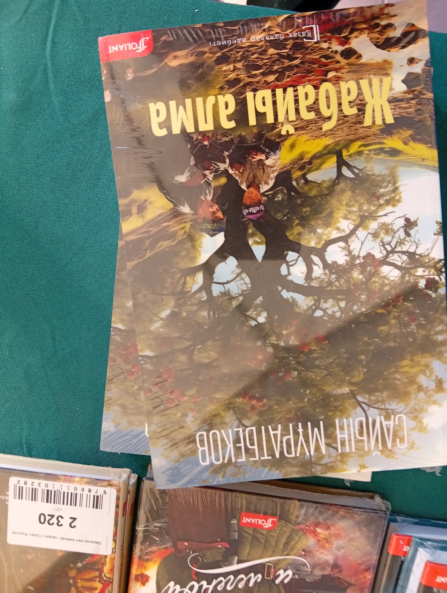 The image shows a close-up, slightly angled view of several books placed on a dark green, textured surface, likely a tablecloth or fabric. The lighting suggests an indoor setting, possibly a bookstore or a home.
The most prominent item is the top book, lying diagonally across the frame, its cover facing upwards and to the right. This book features a bright yellow title in an unfamiliar script, which reads "Жадаи аима" from right to left. Below this title, a stylized image depicts a large tree with red fruits, its branches creating a natural frame for several individuals, appearing to be children or young adults, positioned upside down relative to the tree. One of these individuals, visible on the left, wears a purple head covering with white markings. At the top left corner of this book cover, a red logo with white text reads "FOLIA NT". Above the yellow title, there is a smaller line of text in the same script, "Казак далаы даибметі".
Beneath this top book, another book is partially visible, also with its cover facing upwards. The visible portion of its cover shows the same tree and figures. A portion of a yellow text appears on its cover, which reads "САНБЫН МУРАТБЕКОВ", oriented from right to left.
To the bottom left of the frame, another book is more fully visible, lying horizontally. Its cover displays a more intricate design with what appears to be a military-themed illustration and flames. The text on this cover is also in the unfamiliar script, with a prominent "FOLIA NT" logo in a red square at the top left. A barcode and some text, including the numbers "2 320" and "9 786011 03282", are visible on a sticker on the lower part of this book.
The overall scene is composed of stacked books, suggesting a retail or collection display. No specific landmarks or elements indicating the city of Kostanay, Kazakhstan, are present in the image.