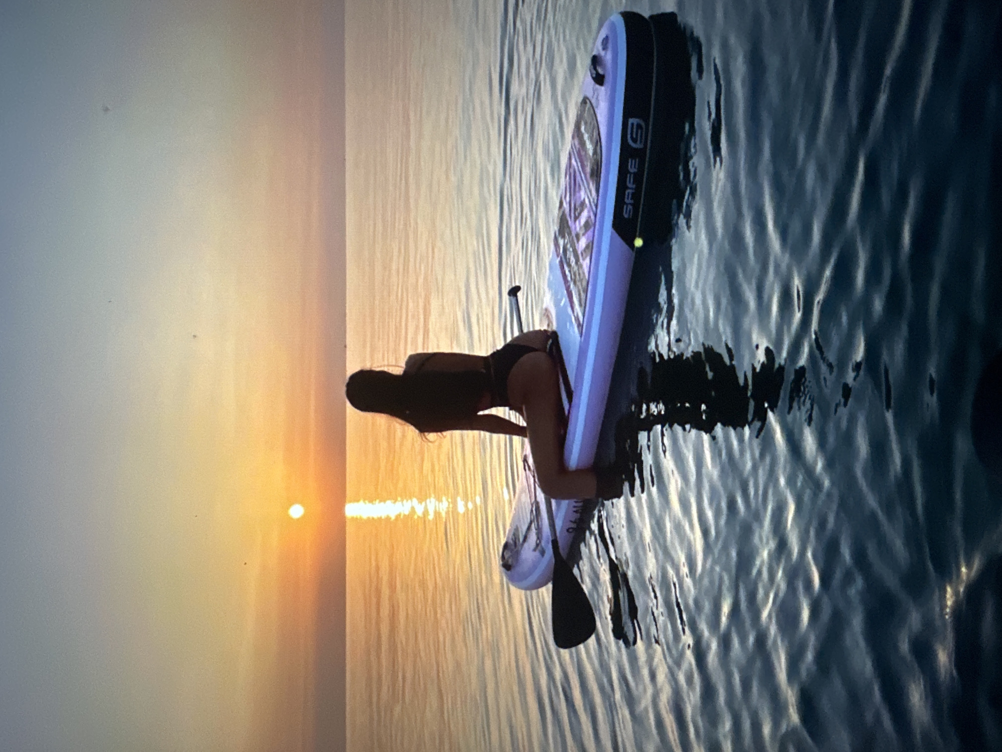 A woman with long, dark hair is sitting on a stand-up paddleboard in calm water during sunset. The sun is low on the horizon, casting a warm glow across the sky and reflecting on the water. The paddleboard is light-colored with black trim and a graphic design on the deck. A paddle rests on the side of the board, and its blade is submerged in the water. The woman is wearing a dark-colored swimsuit. The overall scene depicts a serene and peaceful moment in nature.