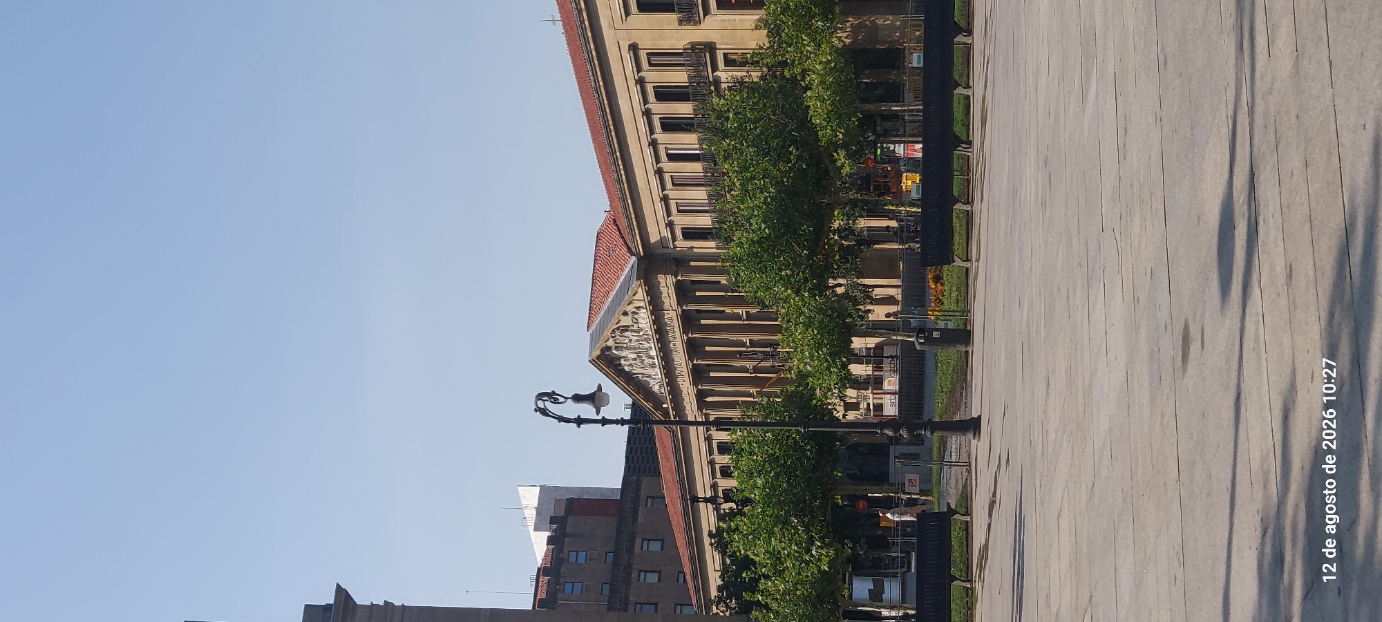 This is a photograph of the Palacio de Navarra in Pamplona, Spain. The building features a neoclassical facade with a prominent pediment adorned with sculptures. A lamppost stands in the foreground, casting a shadow on the paved plaza. Several benches are visible, as is a stand of trees. A person walks in the distance. The sky is clear and blue. The timestamp in the lower left corner reads "12 de agosto de 2026 10:27".