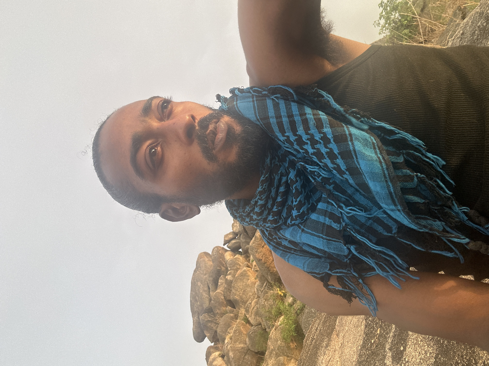 A dark-skinned man is facing the camera, wearing a black tank top and a blue and black patterned scarf. He has a beard and dark hair. Behind him are large, tan-colored rock formations and sparse vegetation against a pale sky. The location is likely near or on one of the rock formations in Jos, Nigeria.