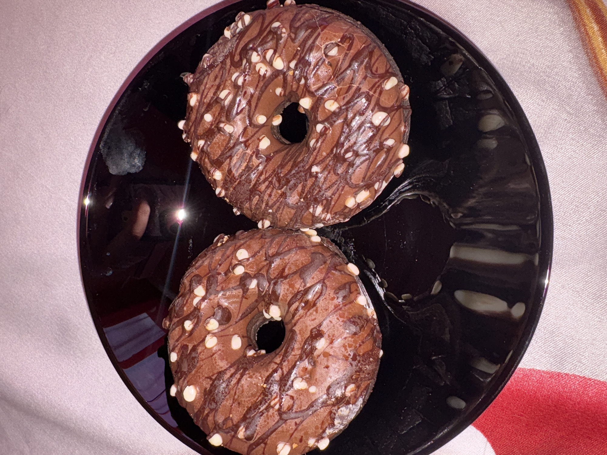 Two chocolate glazed donuts with white chocolate chips and a chocolate drizzle are placed on a glossy black plate. The plate sits on a patterned cloth, which has sections of pink, red, and white. The lighting in the image is dim, creating reflections on the plate. The scene appears to be an indoor setting. There are no people or identifiable landmarks that would indicate a specific location in Vratsa, Bulgaria.