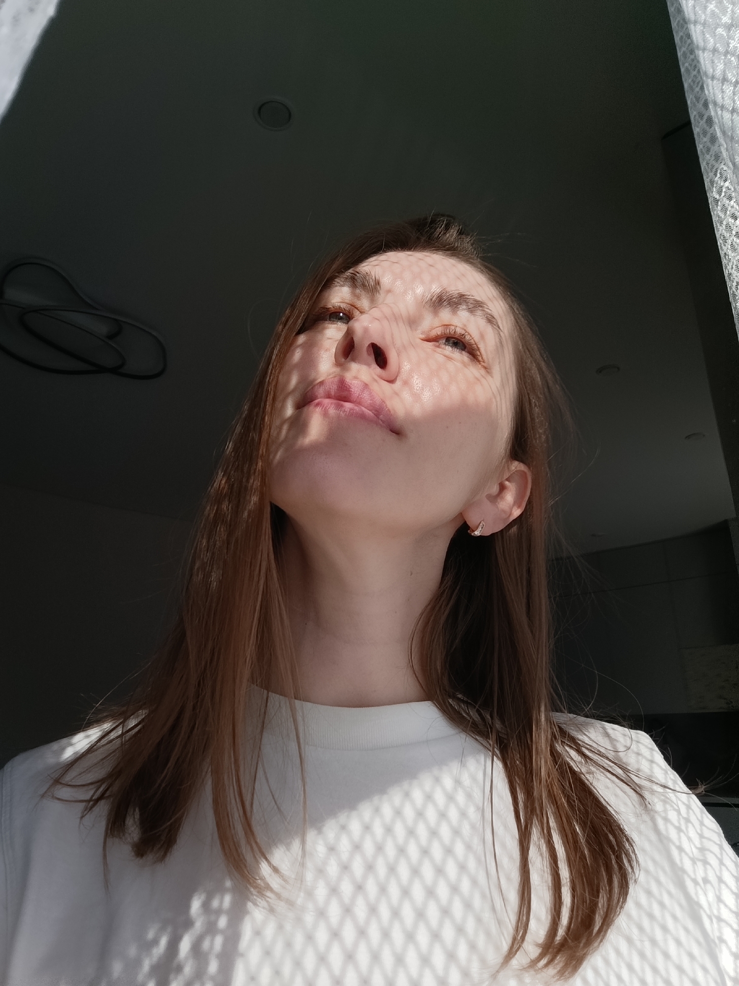 A young woman with light brown hair and fair skin is seen from a low angle, gazing upward. Her face and upper chest are covered in a grid-like pattern of sunlight and shadow, possibly cast by a window covering. She is wearing a plain white t-shirt and small earrings. The background is mostly dark, with a partial view of a ceiling, lighting fixture, and a white, lacy curtain. The image was taken in Chaikovsky, Russia.