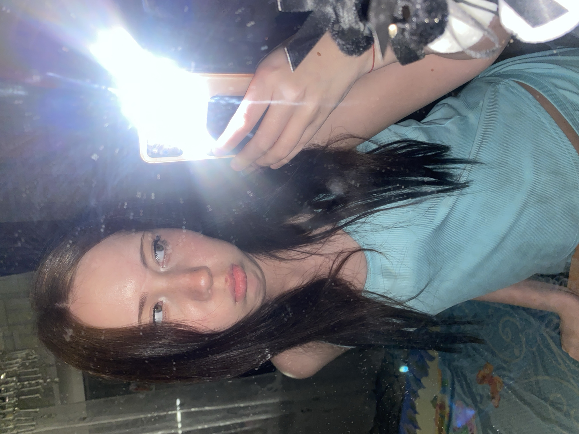 A young woman is taking a selfie with a cell phone. The flash from the cell phone is very bright. She is wearing a light blue shirt and has long, dark hair. The environment is dimly lit and appears to be indoors. The location is unknown.
