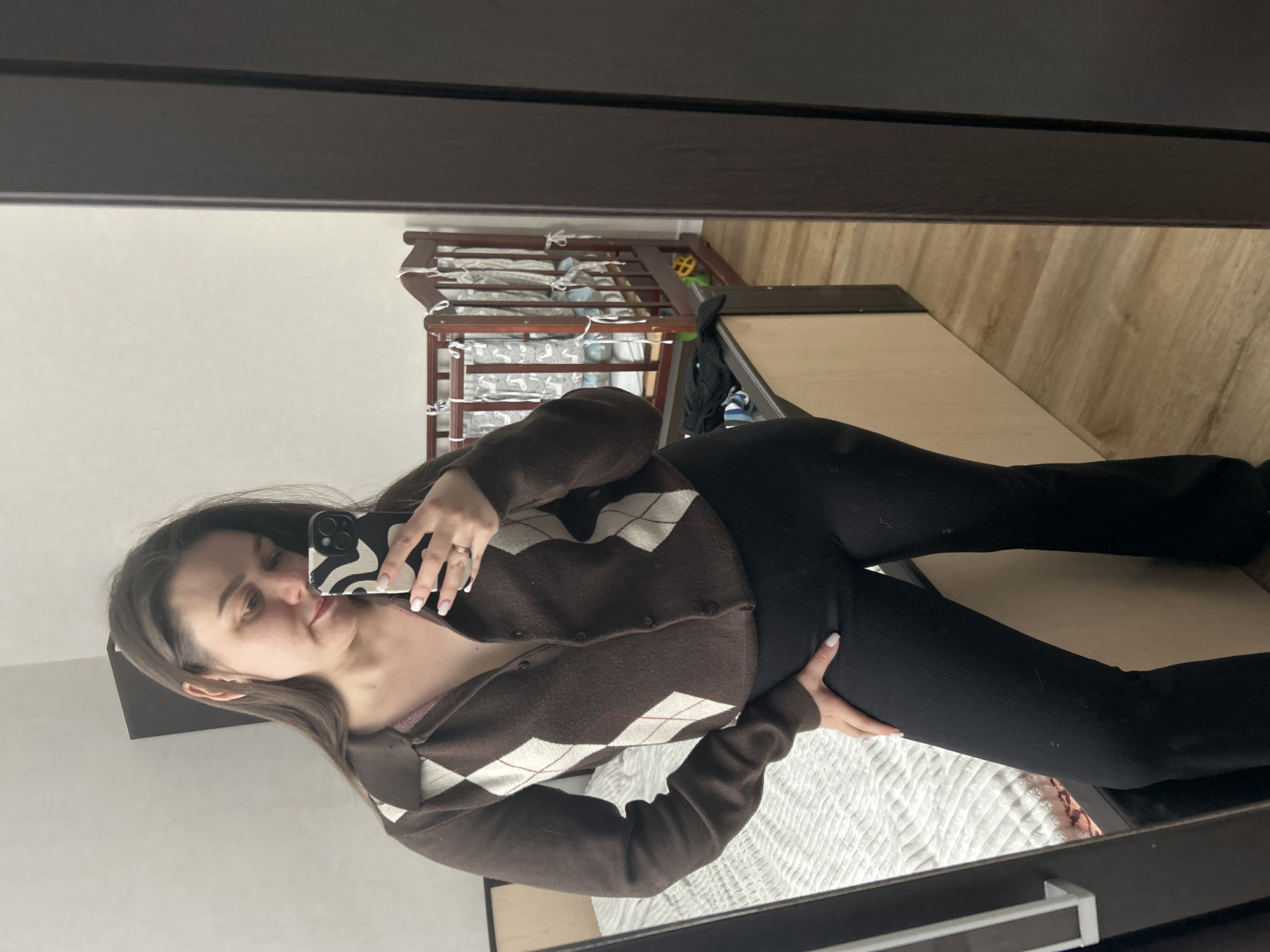 A woman is taking a selfie in a mirror. She is wearing a brown and white argyle sweater and black pants. In the background, there is a crib and a wooden floor. The location is a domestic interior, but the city of Chaikovsky, Russia cannot be verified.