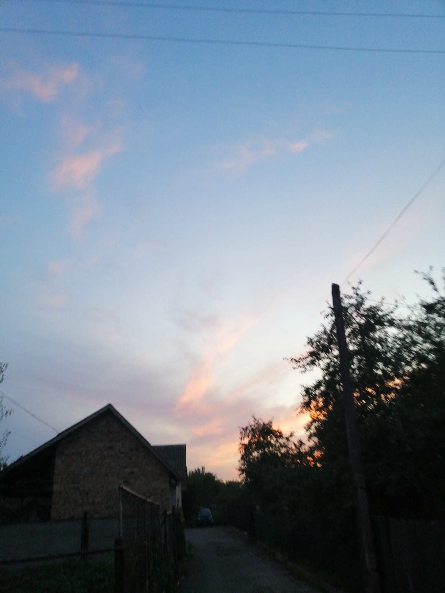 A rural scene unfolds under a twilight sky in Lanivtsi, Ukraine. The sky transitions from a clear blue at the top to soft pink and orange hues near the horizon, indicating dusk. A series of dark power lines crisscross the upper portion of the frame. In the foreground, a stone building with a dark, sloping roof is visible on the left. A chain-link fence runs along the front of the property. To the right, a dark, leafy tree partially obscures a utility pole. Beyond the building and fence, a dark asphalt road leads into the distance, with more trees and foliage lining the right side of the frame. A dark-colored car is parked on the side of the road to the right of the building. The overall impression is of a quiet evening in a residential area, likely on the outskirts of town.