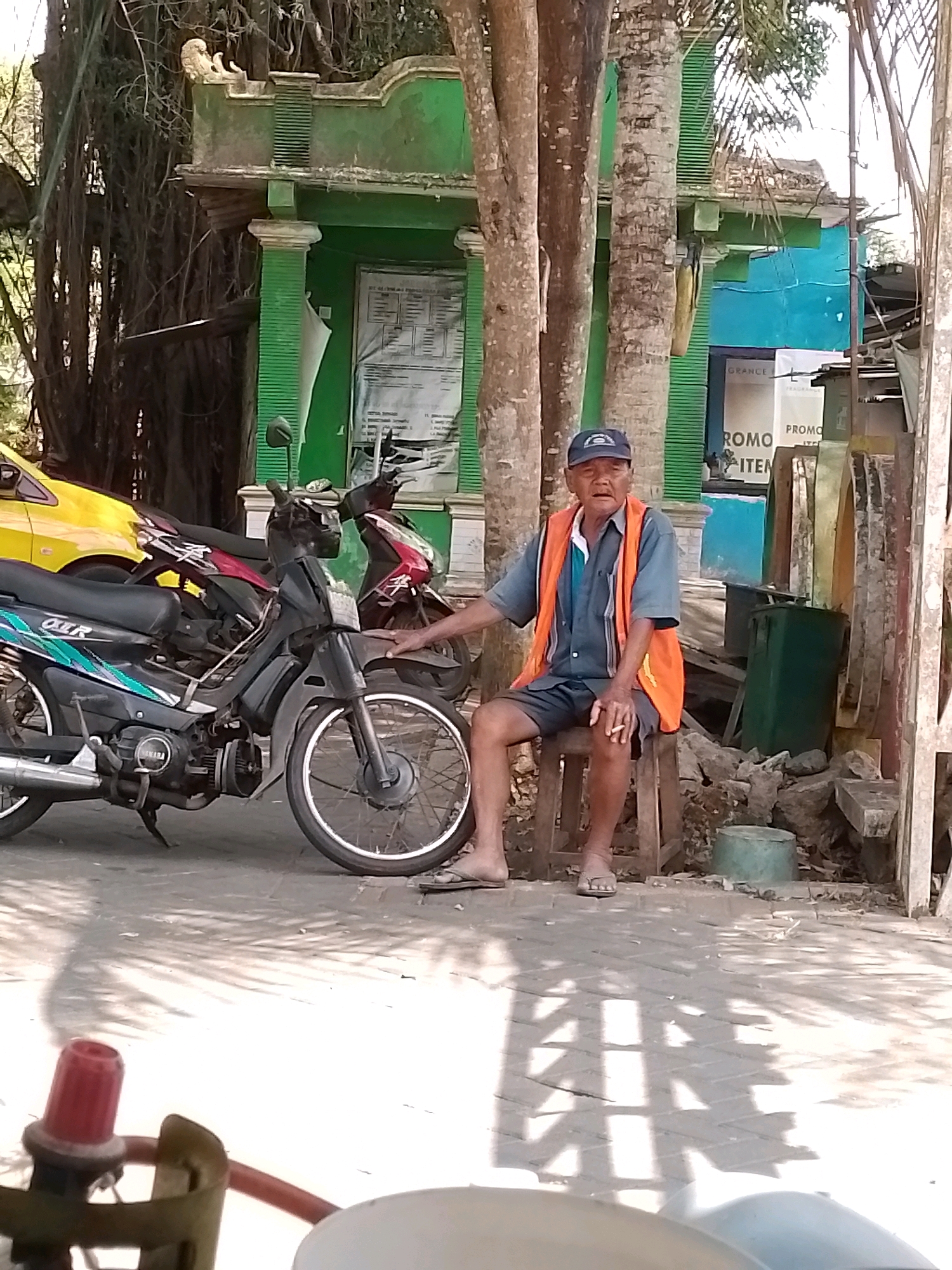 An elderly man in a blue shirt, grey shorts, and an orange safety vest sits on a wooden stool in front of a green building in Wonosari, Indonesia. He is wearing a blue baseball cap and appears to be smoking a cigarette. He has his left arm resting on his leg, and his right hand holds the cigarette. In the background, there are trees, a yellow car, and two motorcycles. The ground is paved with interlocking bricks, and shadows are cast on it from overhead foliage. A red regulator is visible in the foreground. The building appears to be a small commercial establishment with signage that includes "GRANCE" and "PROMO".