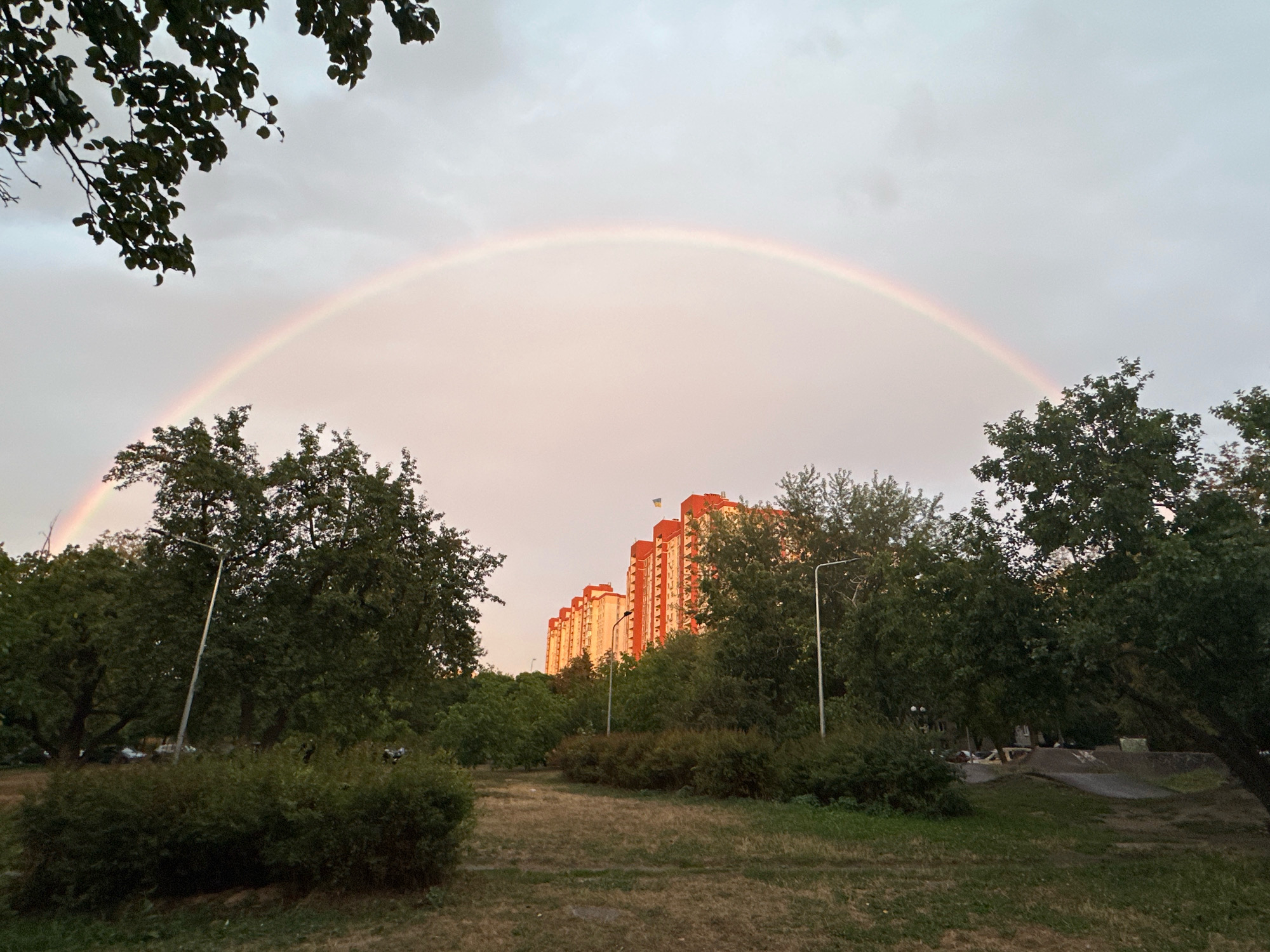 The image depicts a cityscape with a prominent rainbow arcing across a cloudy sky over Kyiv, Ukraine. In the foreground, a grassy area is dotted with bushes and trees, leading towards a park-like setting with ramps and structures that appear to be part of a skate park. Beyond the park, a row of tall, orange-and-white apartment buildings stretches horizontally, with a Ukrainian flag visible atop the tallest building. The scene is captured during the day, likely after a rain shower, given the presence of the rainbow. The overall impression is one of urban nature bathed in the soft light of a post-rain sky.