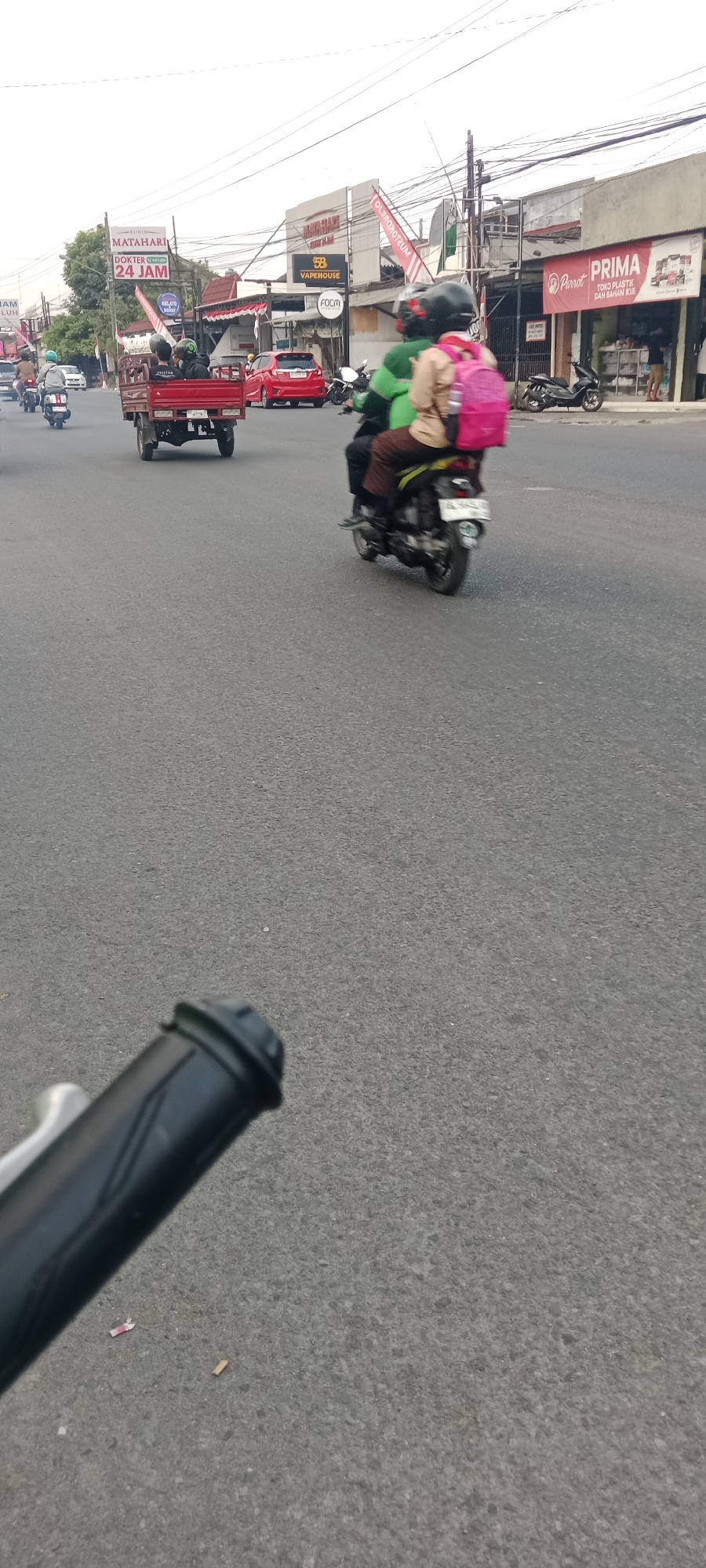 In Maguwoharjo, Indonesia, a street scene unfolds with several vehicles and pedestrians. A red, three-wheeled truck is moving forward, carrying passengers in its rear. To its right, a motorcycle is also moving, with two people on it, one wearing a pink backpack. Further ahead, a red car is visible. On the side of the road, various shops and signage are present, including "Matahari Dokter 24 Jam," "Vapehouse," "Foom," and "Prima Parrot Toko Plastik dan Bahan Kue." The road surface is asphalt, and power lines are visible overhead. The image appears to be taken from a moving vehicle, possibly a bicycle or another motorcycle, as evidenced by the handlebars and a cigarette butt in the foreground. The sky is overcast.