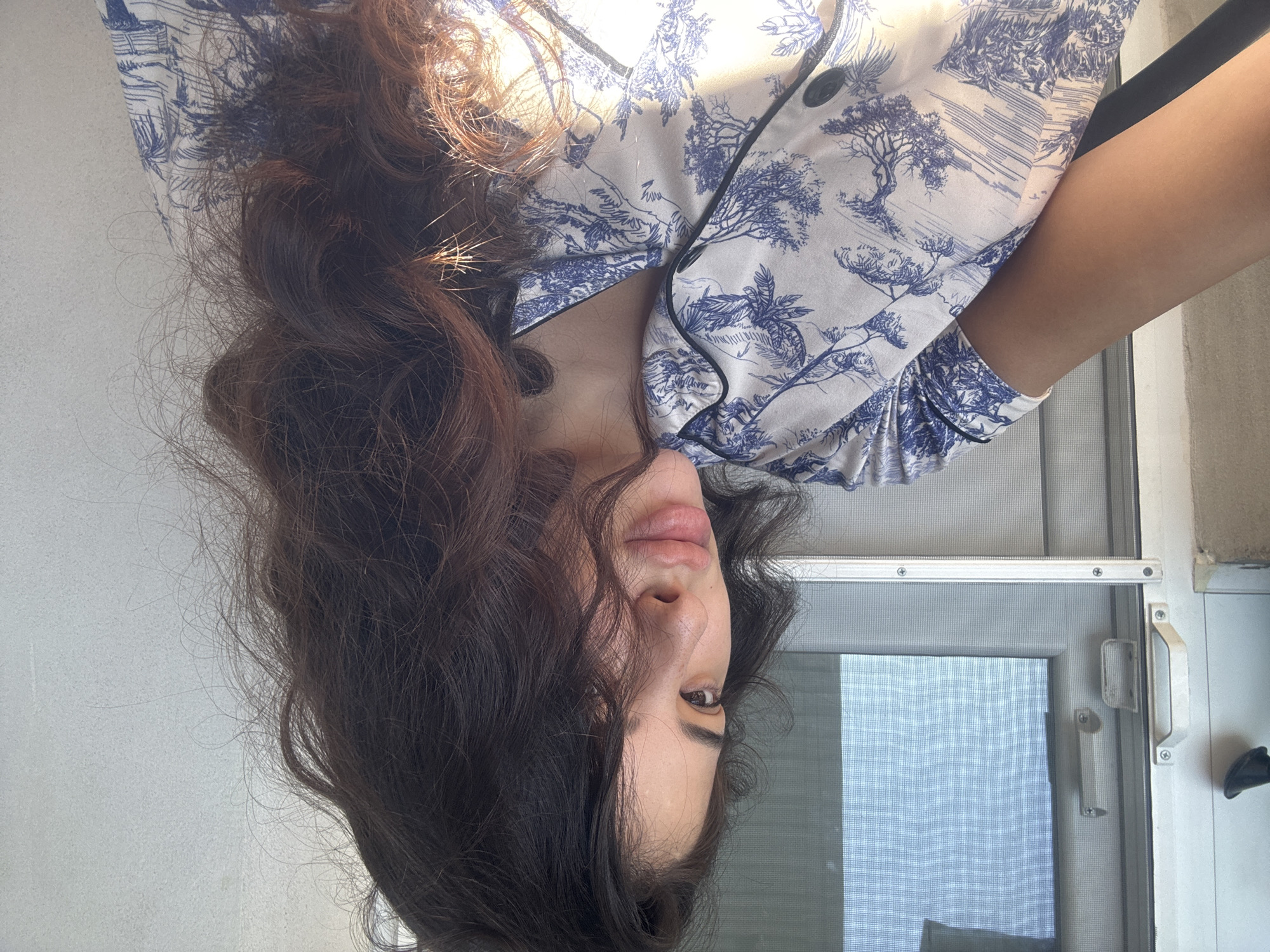 The image shows a close-up, head-and-shoulders shot of a young woman with long, dark, wavy hair, looking towards the right of the frame. She is wearing a white pajama top with a blue toile-like pattern of trees and landscapes. Her face is partially obscured by her hair. The background suggests an indoor setting near a window with a screen, possibly on a balcony or a room with a sliding door. The lighting indicates it is daytime, with sunlight casting highlights on her hair and clothing. There is no information in the image to suggest a location in Plovdiv, Bulgaria.
