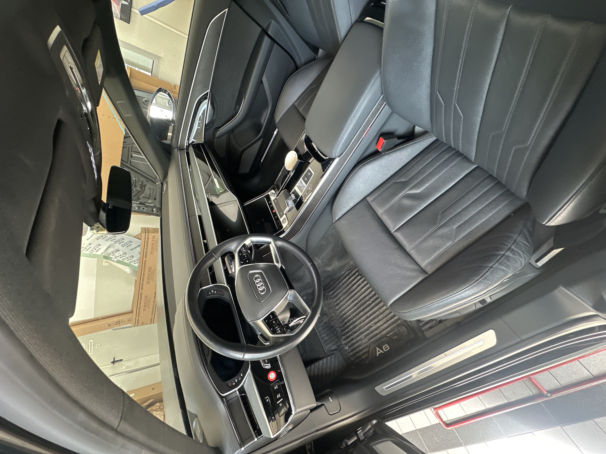 The image shows the interior of a black Audi A8, viewed from the front passenger seat looking towards the driver's side. The car's dashboard, steering wheel, center console, and front seats are visible. The dashboard features multiple screens and modern controls. The steering wheel is black leather with silver accents. The center console has a gear shifter, cupholders, and other buttons. Both front seats are upholstered in black leather with intricate stitching. A floor mat with the "A8" logo is visible on the driver's side floor. The car appears to be parked indoors, possibly in a garage or workshop, as indicated by the presence of boxes and what looks like garage equipment in the background visible through the windshield. The lighting suggests it is daytime. There are no people in the image.