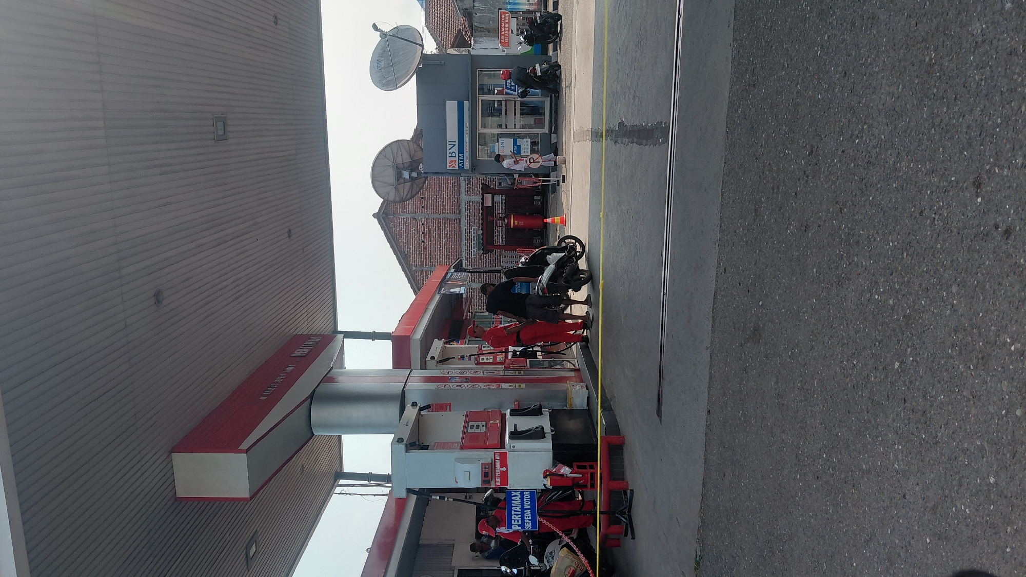 The image depicts a gas station in Rejasari, Indonesia. A Pertamina gas pump is visible on the left, with a sign indicating "PERTAMAX SEPEDA MOTOR." Two attendants in red uniforms are present near the pumps. One is holding a nozzle, and the other is standing next to him. A man in a black shirt is standing near a scooter. In the background, there is a building with BNI ATM signage and two satellite dishes mounted on the roof. Several motorbikes are parked around the gas station. The ground is paved with asphalt and concrete. The overall scene appears to be an active, everyday operation at the gas station.
