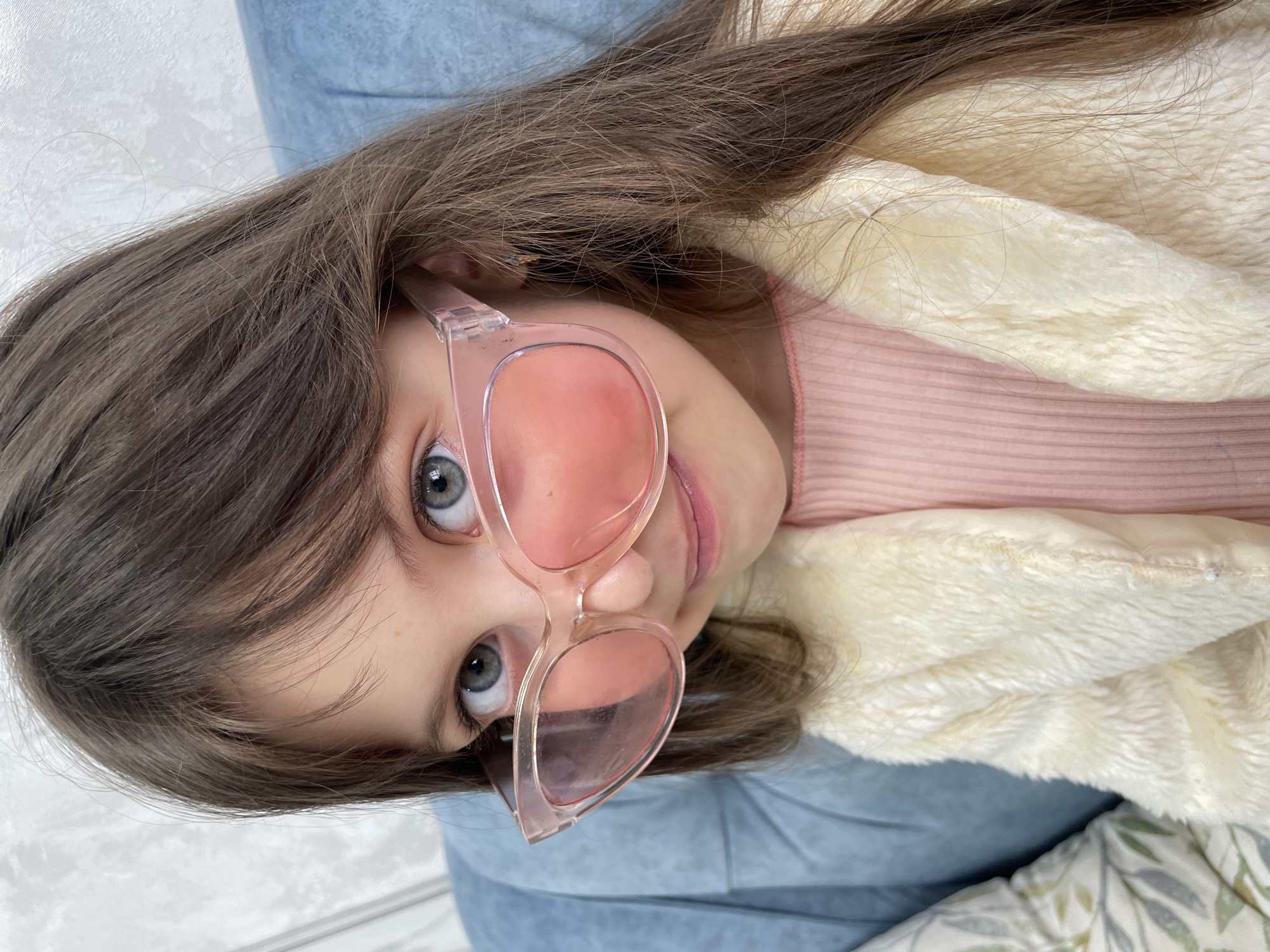 A young girl with long brown hair is shown at a tilted, close-up angle, wearing pink-tinted glasses. She has light skin and grey-blue eyes. She is wearing a light pink shirt and a cream-colored, plush jacket or blanket. She appears to be lying on a light blue surface, possibly a couch or bed. A pillow or blanket with a leaf pattern is partially visible in the lower right corner. There is no identifiable information in the image to indicate it was taken in Izhevsk, Russia.