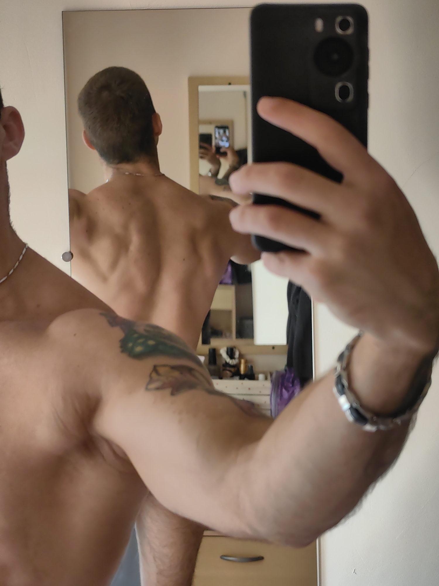The image shows a shirtless person taking a mirror selfie. The person is standing in a room with a large mirror reflecting their back. They have short brown hair, a necklace, and tattoos on their left arm. The room appears to be a bedroom or dressing area, with shelves and furniture visible in the background. The lighting is bright, suggesting daytime. There is no identifiable information in the image that would suggest a specific location in Varna, Bulgaria.