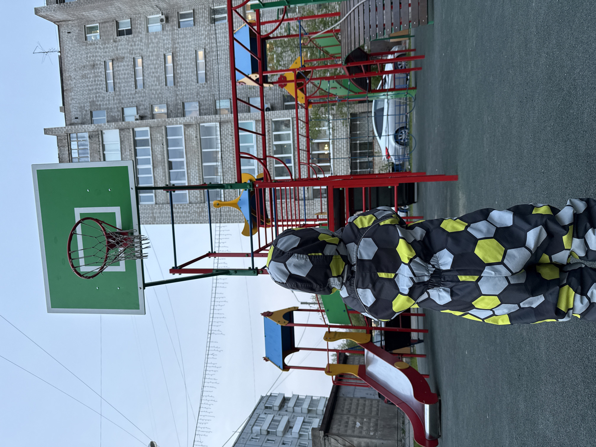 A child wearing a soccer ball pattern jumpsuit is standing on a playground. The playground includes a basketball hoop with a green backboard, a red slide with yellow accents, and a red and blue climbing structure. In the background, a tall apartment building made of grey brick with many windows rises into the sky. There are decorative lights strung overhead. A white car is partially visible in the background. The playground is situated on a green surface. The location is Krasnoyarsk, Russia.