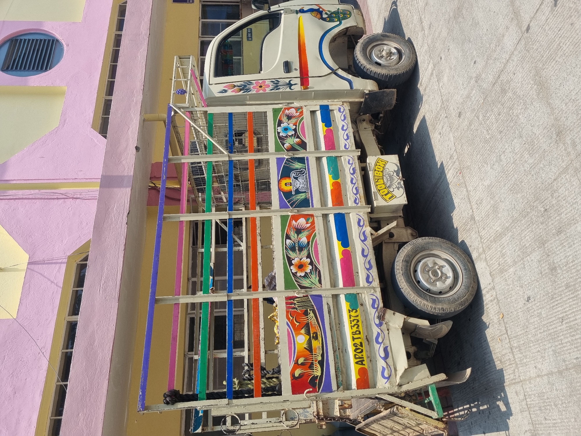 The image displays a cream-colored light commercial vehicle (LCV), specifically a mini-truck, parked on a textured concrete surface under bright daylight in Puttaparthi, India. The vehicle's open cargo bed is surrounded by a metal cage featuring numerous horizontal bars painted in vibrant colors, including purple, pink, green, blue, and orange. The side panels of the cargo bed are extensively decorated with hand-painted artwork, depicting a landscape with a setting or rising sun over mountains, trees, and a boat, alongside intricate floral patterns and a sacred pot (kalasha) design. The license plate "AP02 TB 3373" is visible on the lower left section of the cargo bed. A "TOOLBOX" with a floral design is mounted below the cargo bed. The cabin door features additional painted floral motifs and a peacock design. A coiled dark rope is secured to the front left of the cargo cage, and a small purple ornament hangs from the roof rack above the cabin. The vehicle's wheels, including a visible front right and part of a rear wheel, are on the concrete. In the background, a large, multi-story building is visible, painted in distinct sections of light pink and yellow. This building features multiple rectangular windows and a prominent circular louvered vent on its upper pink section. No individuals are present in the scene, and no actions or interactions are depicted.