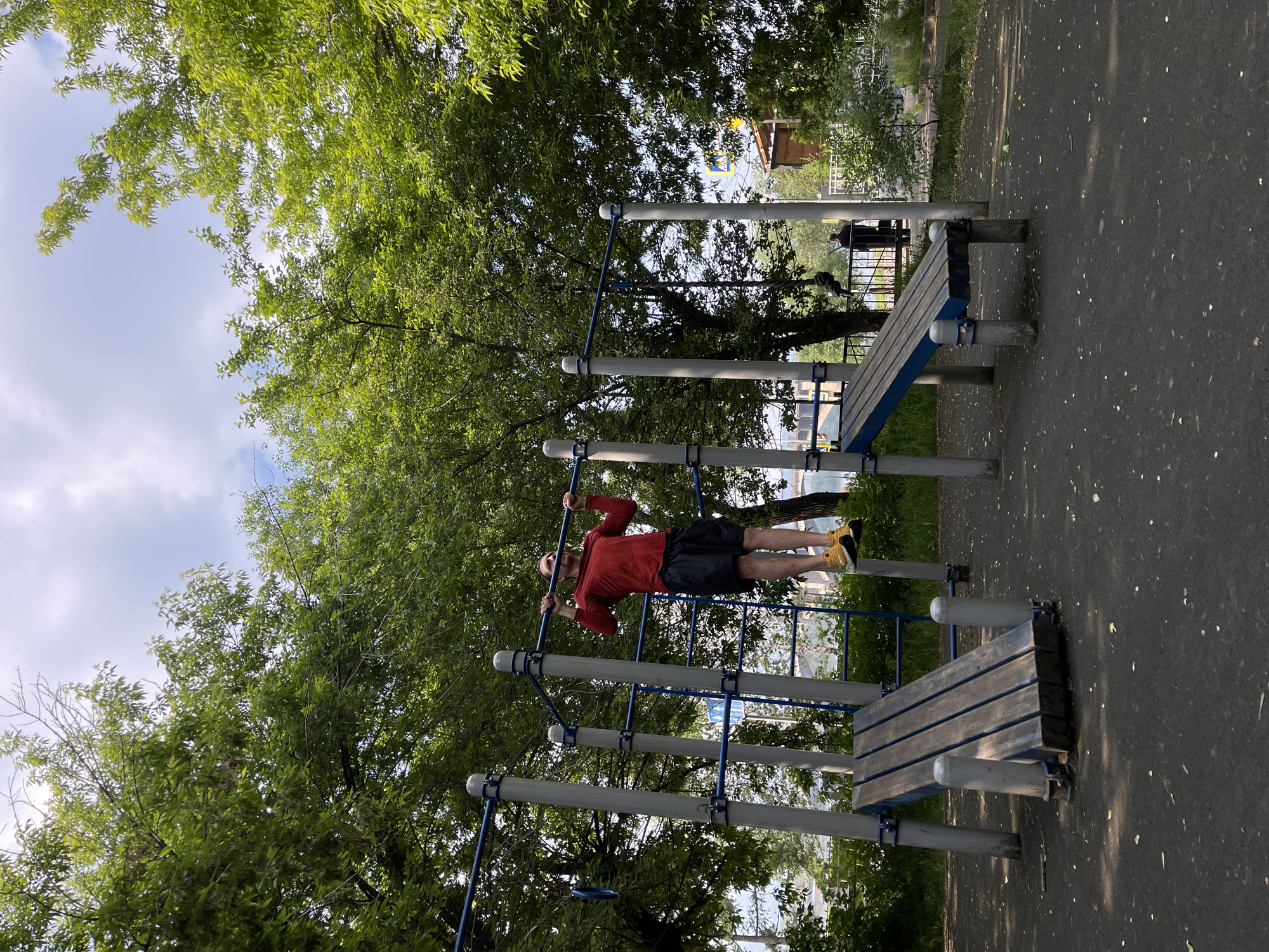 A man is performing a pull-up on a horizontal bar at an outdoor gym. He is wearing a red long-sleeved shirt, black shorts, and yellow sneakers. The gym equipment is blue and gray, with wooden benches. The ground is covered in a black rubberized surface with scattered leaves. Lush green trees surround the gym. In the background, there is a glimpse of a bridge, a body of water, and a building with a yellow and blue flag. The scene is set in Krasnoyarsk, Russia, evident from the flag.