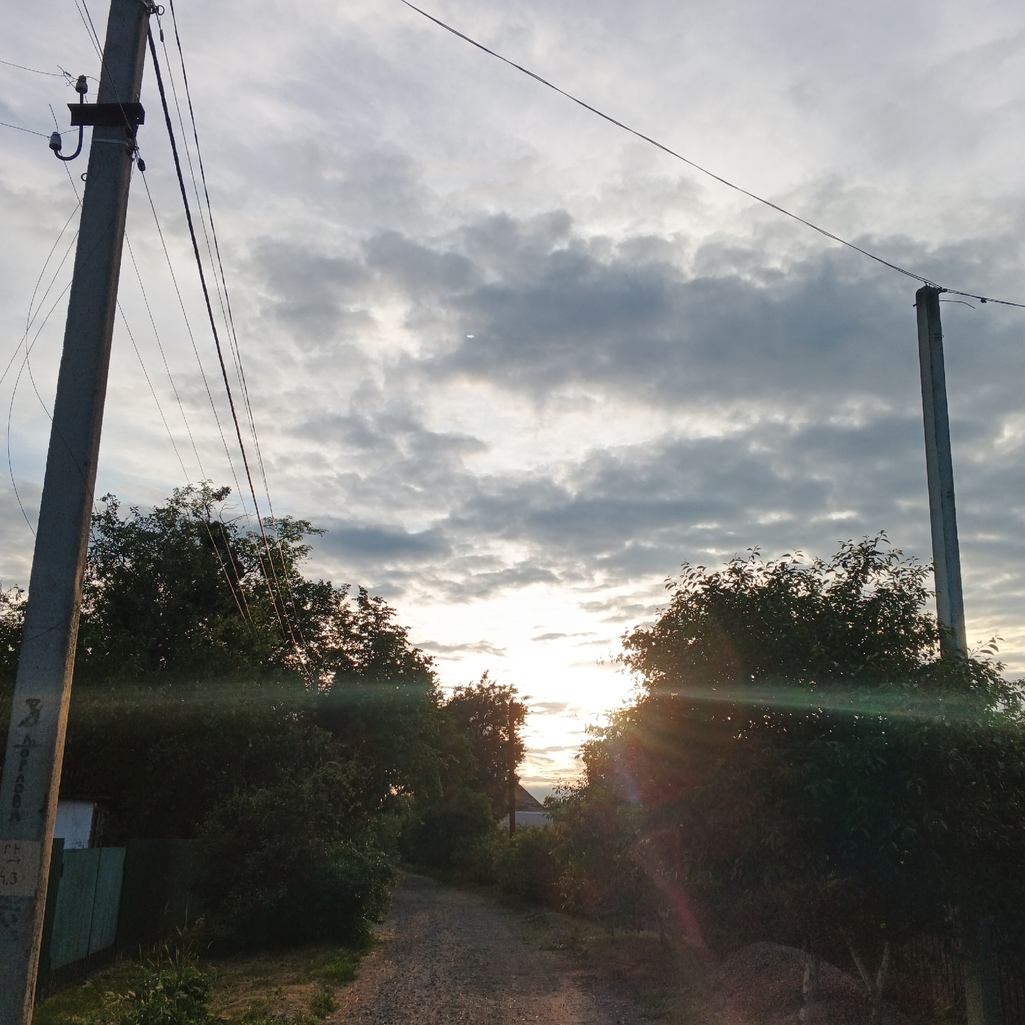 The image depicts a dirt road leading through a wooded area with two utility poles. The sun is setting or rising, casting a warm glow through the trees and clouds. The sky is filled with dramatic, gray clouds. One utility pole is on the left side of the frame, a concrete structure with insulators and wires. The second utility pole is on the right side, also concrete, with wires extending across the scene. Dense green trees line both sides of the road. In the background, a small house with a visible roof can be seen through a gap in the trees. The overall impression is a rural or suburban setting, likely on the outskirts of Smila, Ukraine, at dusk or dawn. There are no people visible in the image.